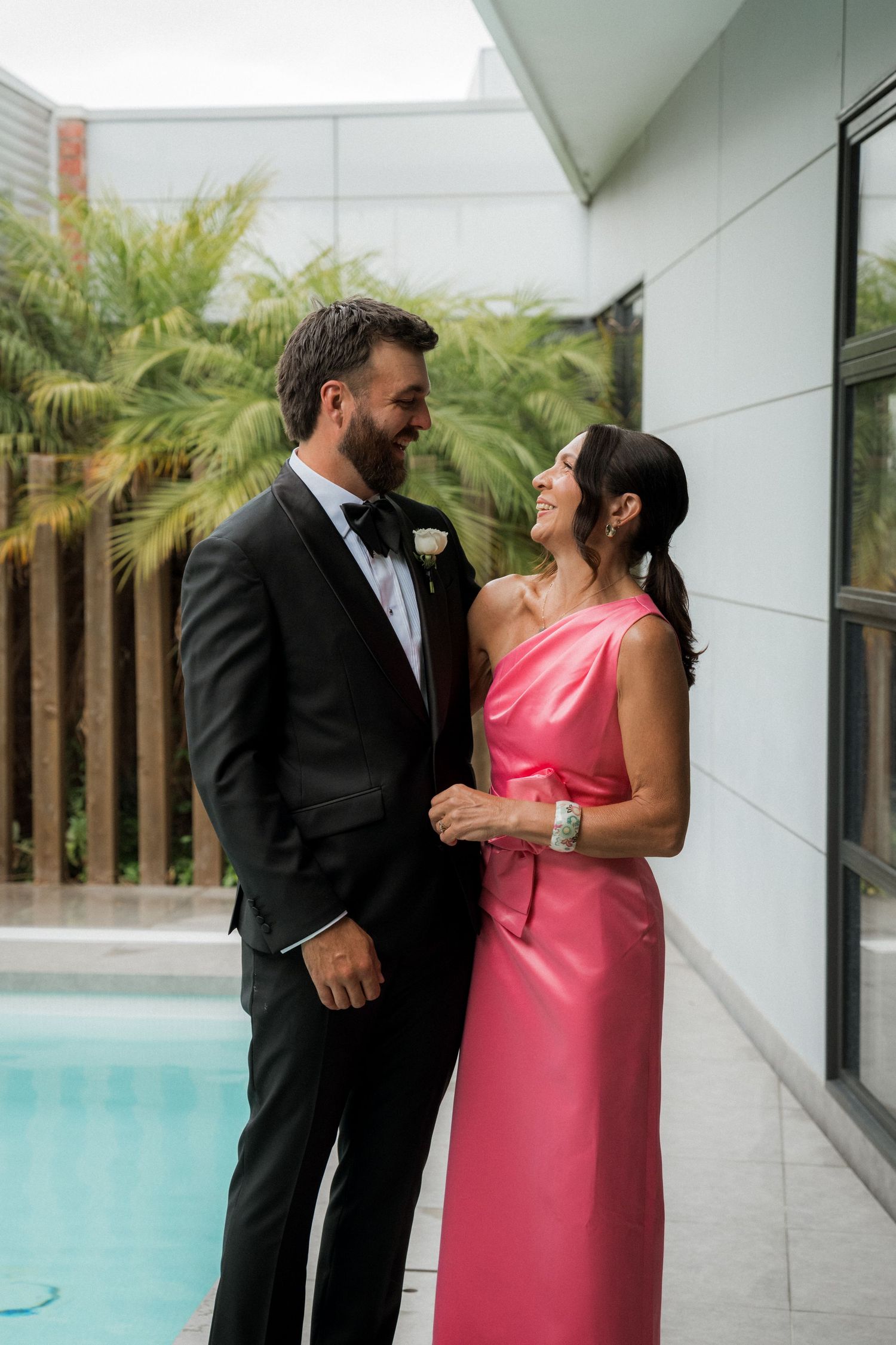 Couple in formal wedding attire smiling at each other beside a pool with tropical greenery backdrop.