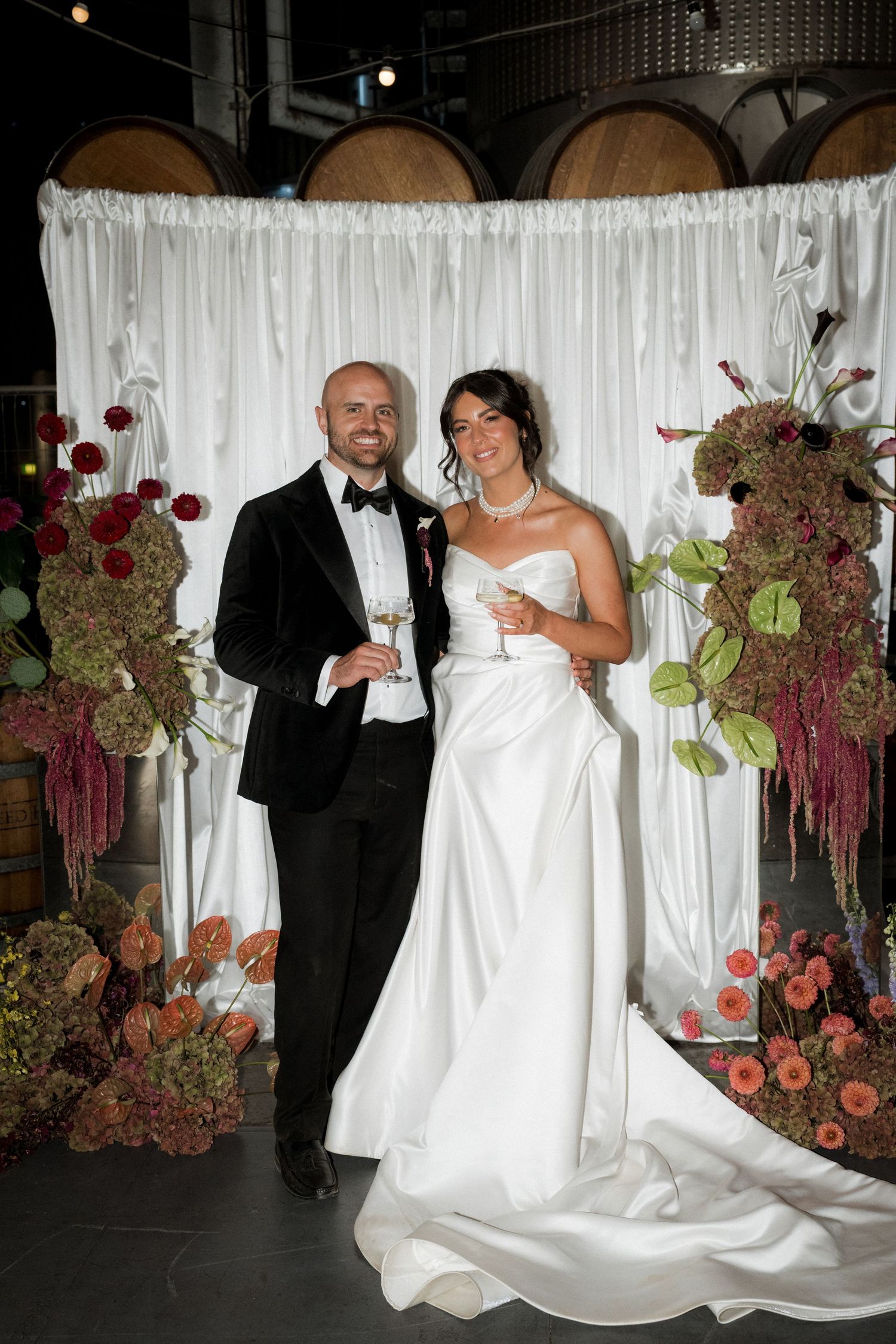 Wedding couple poses in front of white backdrop with elegant burgundy floral arrangements.