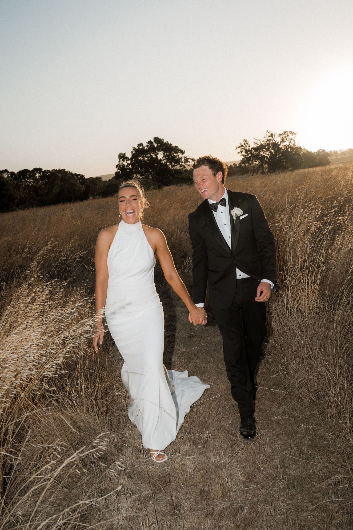 A couple walks hand in hand through golden grass at sunset in their wedding attire.