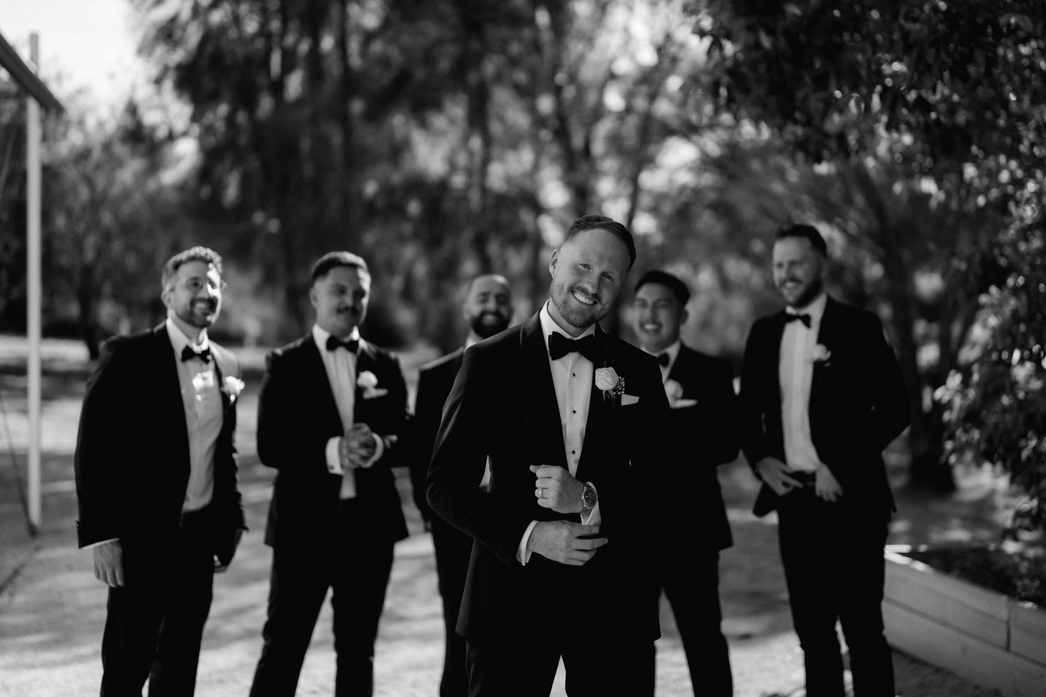 Wedding party members in formal attire gather outside for a casual group photo in black and white.