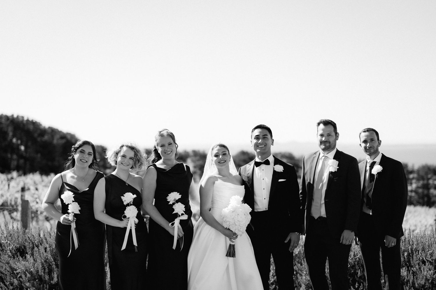 A wedding party in black and white poses together outdoors with bridesmaids in dark dresses.