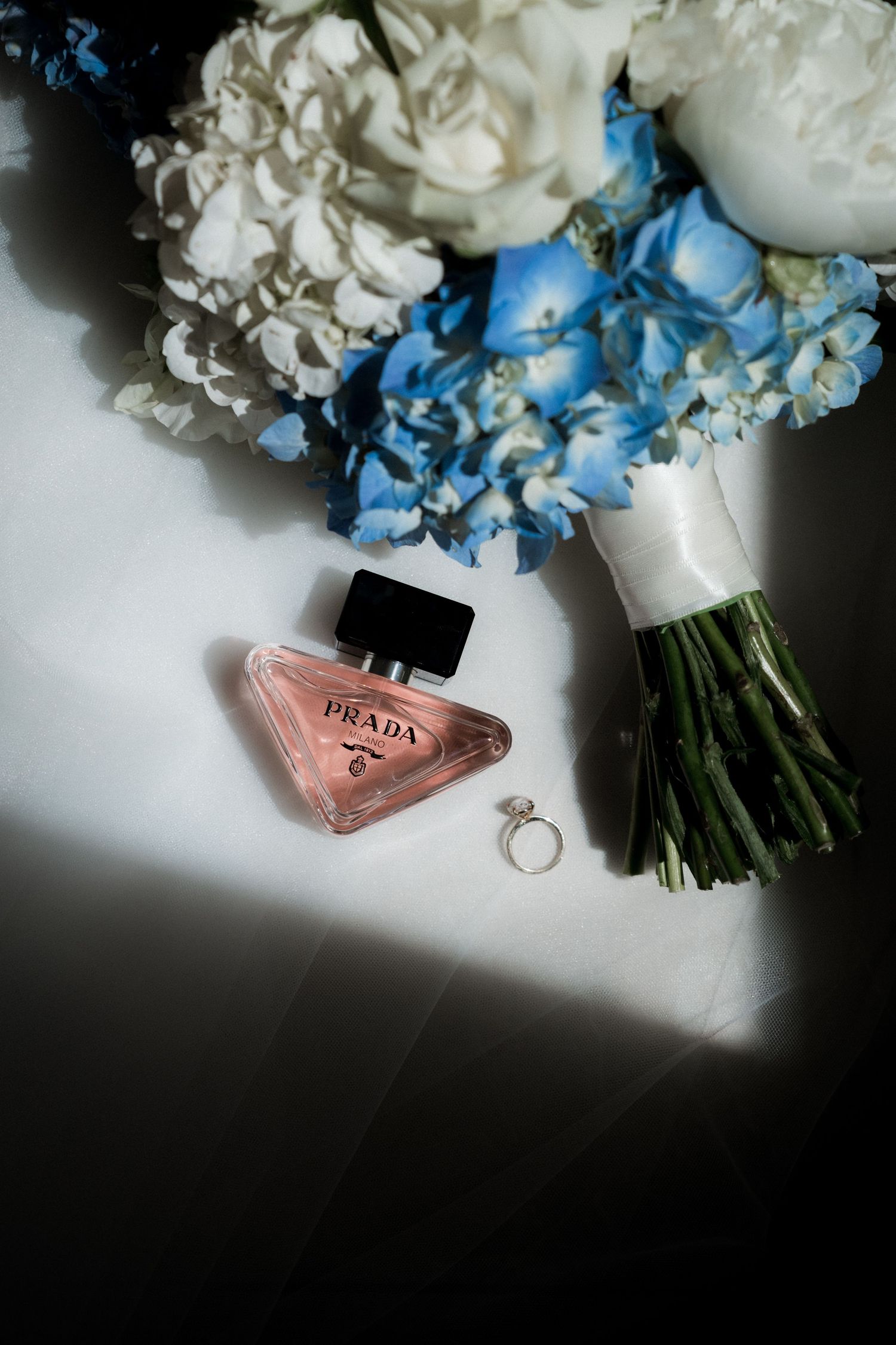 Wedding bouquet of white and blue flowers with a pink Prada perfume bottle and ring on white surface.