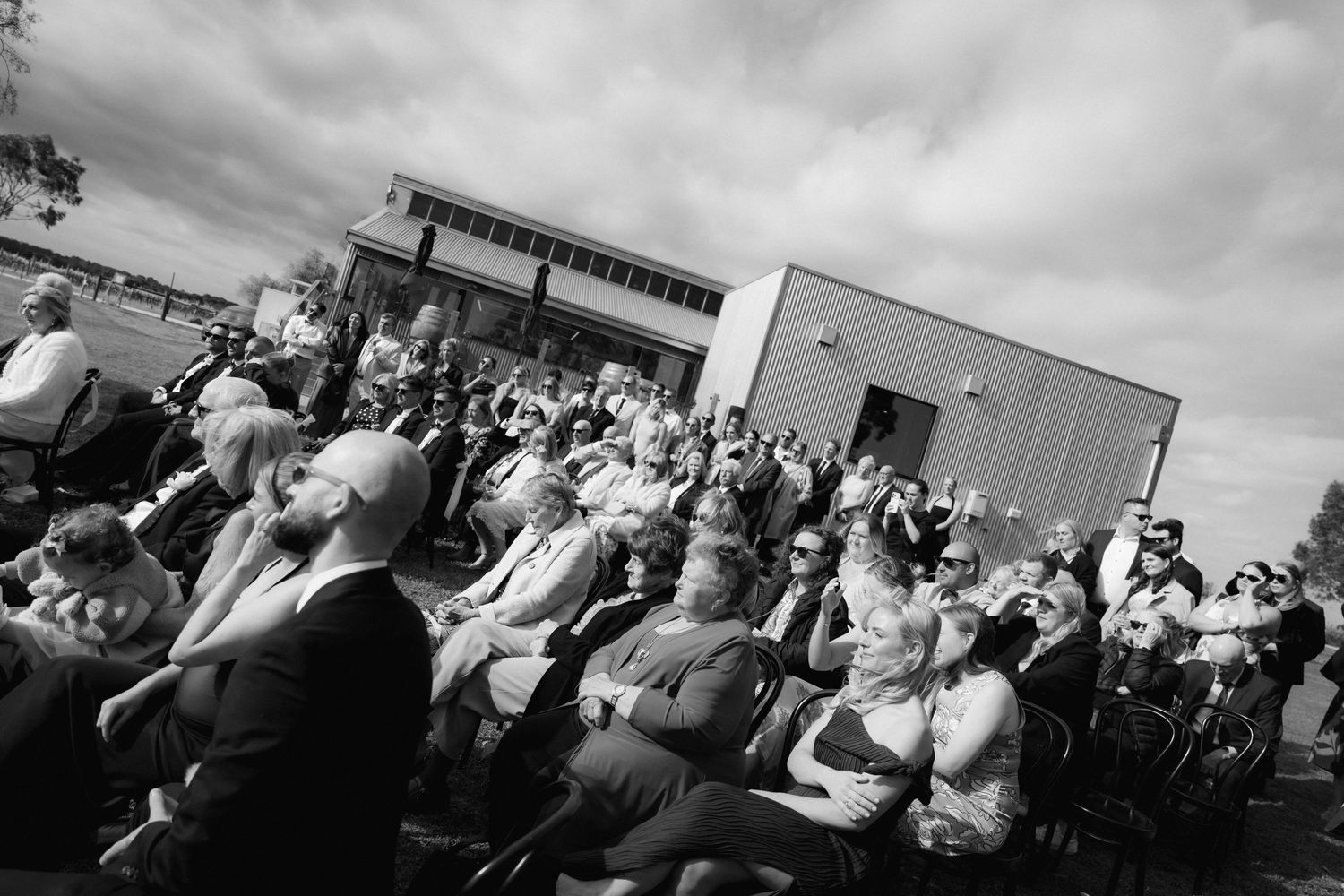 Large crowd of wedding guests gathered outdoors under cloudy skies for ceremony.