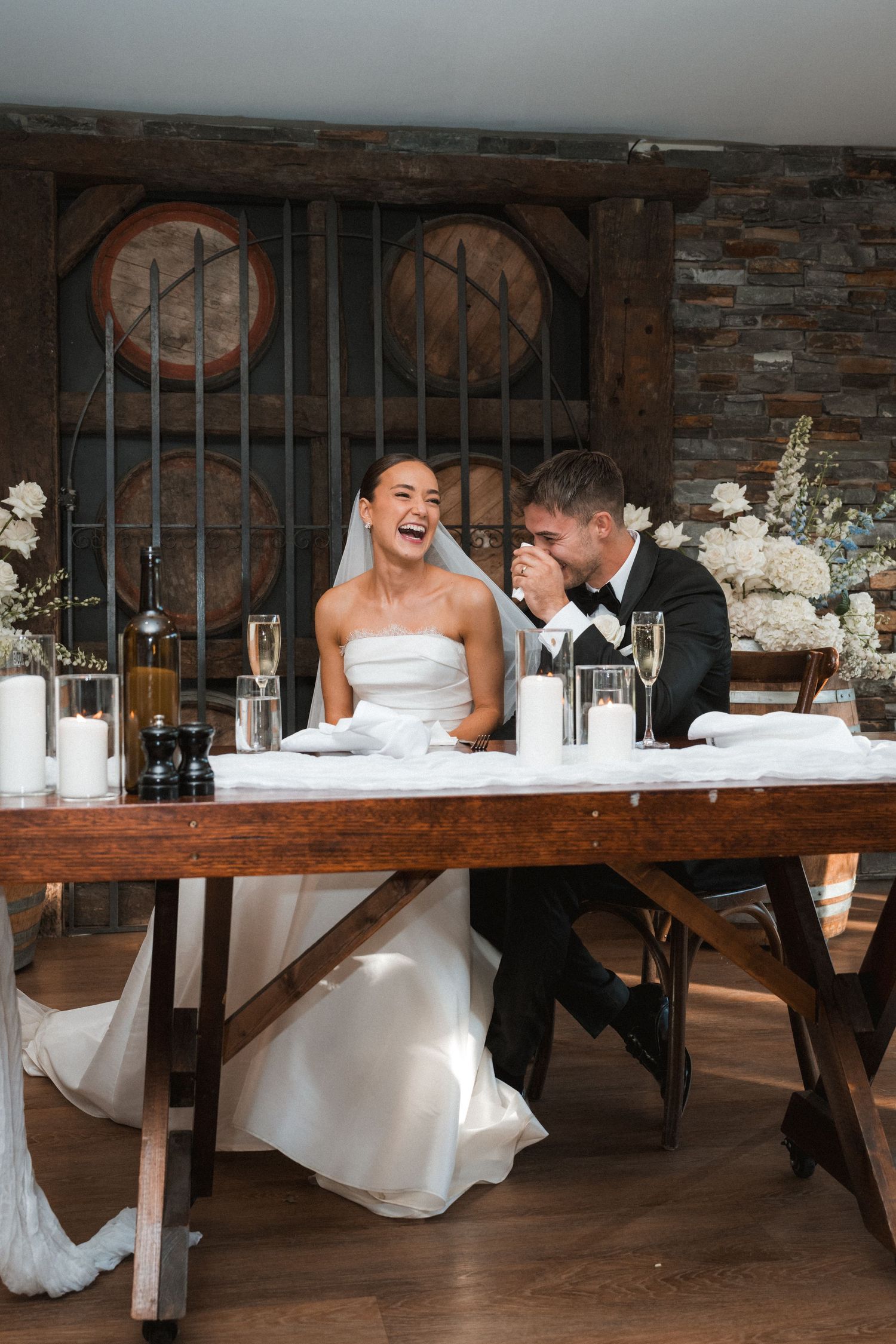 Wedding reception setup with couple seated at rustic wooden head table with white floral decorations.