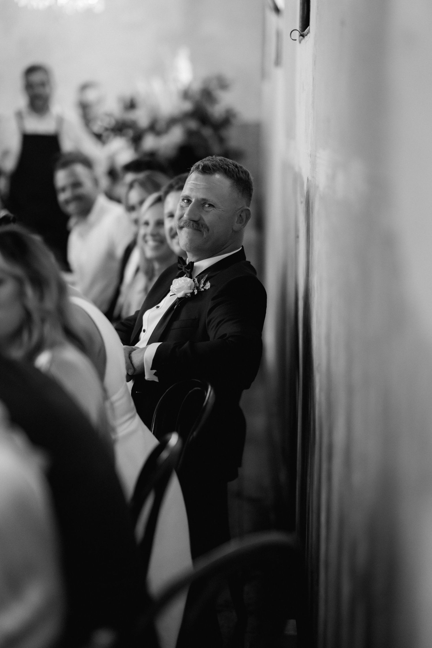 Black and white candid photo of a guest in formal attire leaning against wall during a wedding celebration.