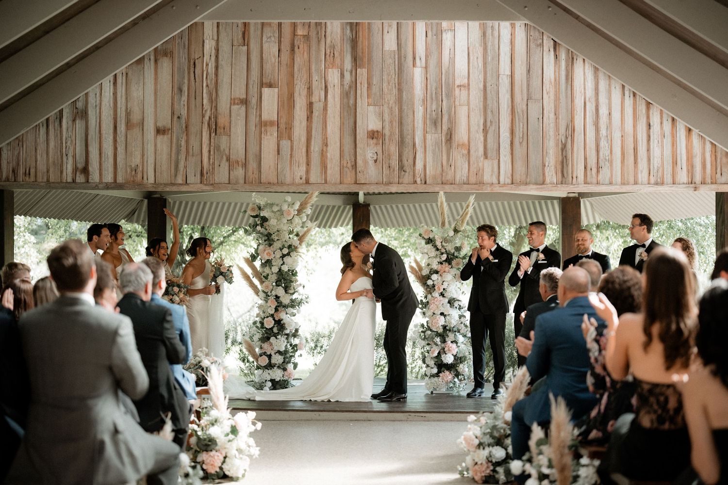 Wedding party dances and celebrates inside a rustic wooden barn venue decorated with white floral arrangements.