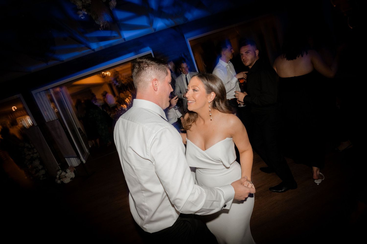 Dancing silhouettes move across a dark blue-lit dance floor at an evening celebration.