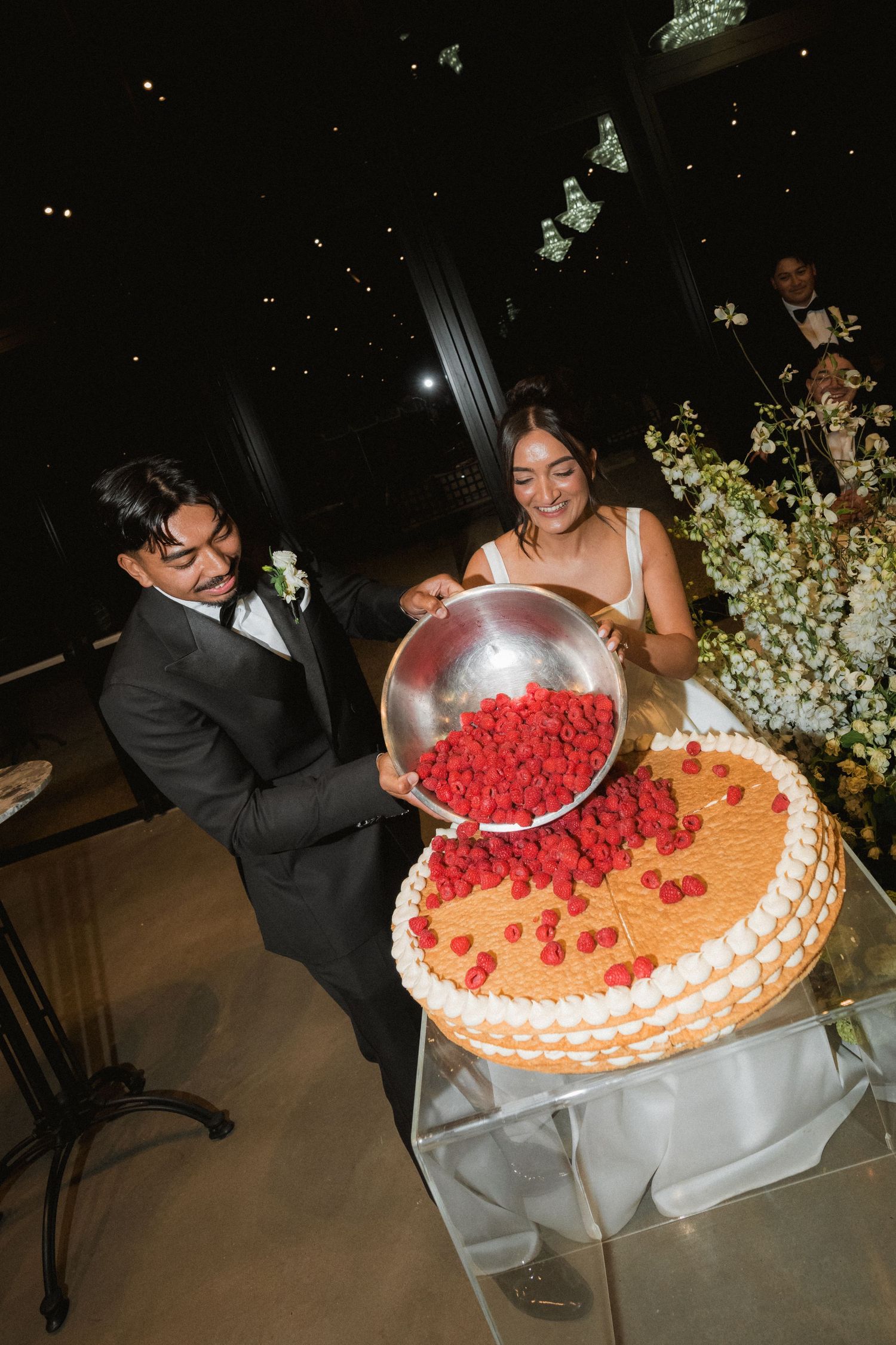 A groom in a dark suit pours fresh raspberries from a metal bowl onto a wedding cake as the smiling bride watches.