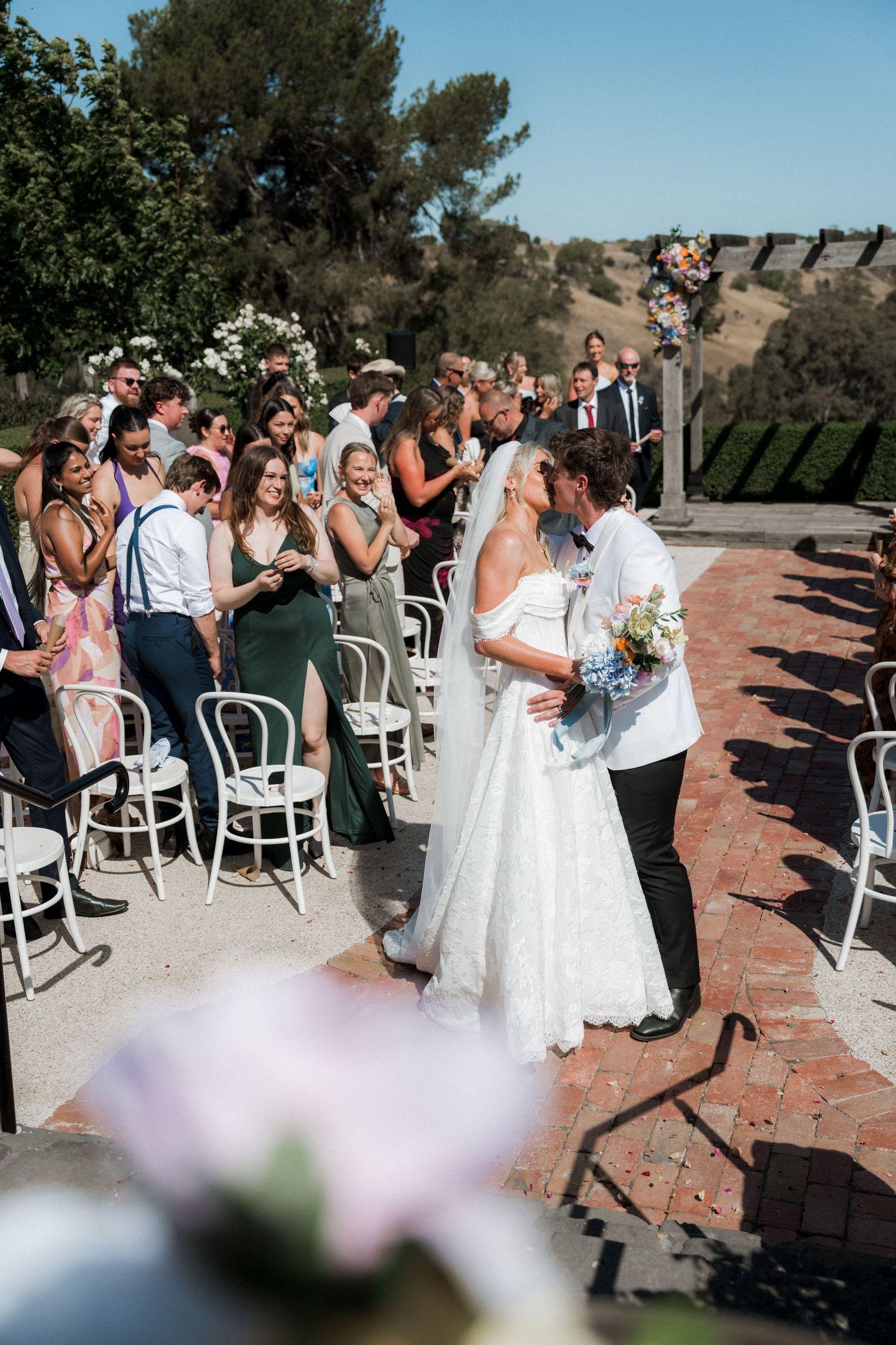 Bride and groom share a kiss at outdoor wedding ceremony while guests applaud along the aisle.