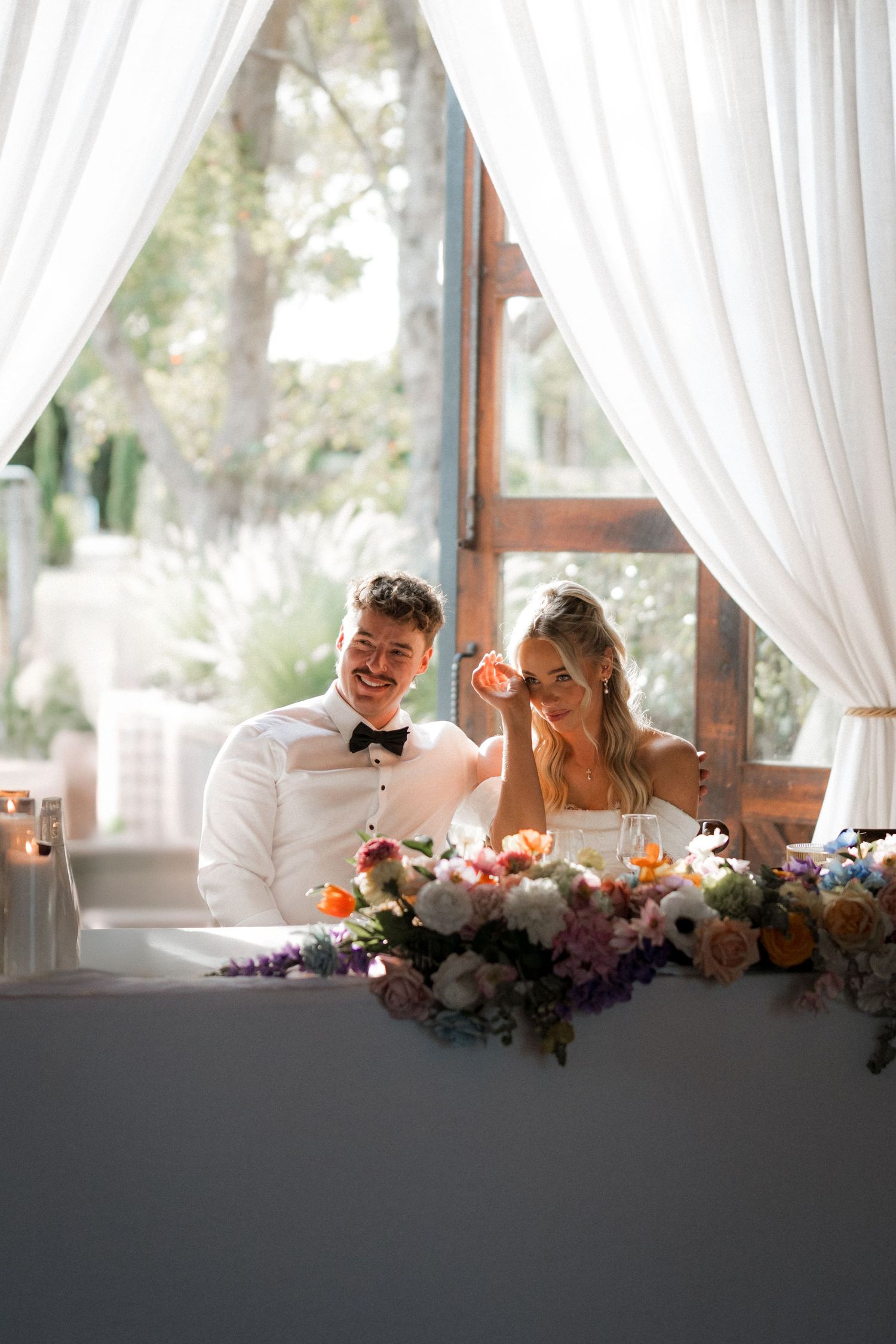 A wedding couple shares a tender moment at their reception table decorated with floral arrangements and white draping.