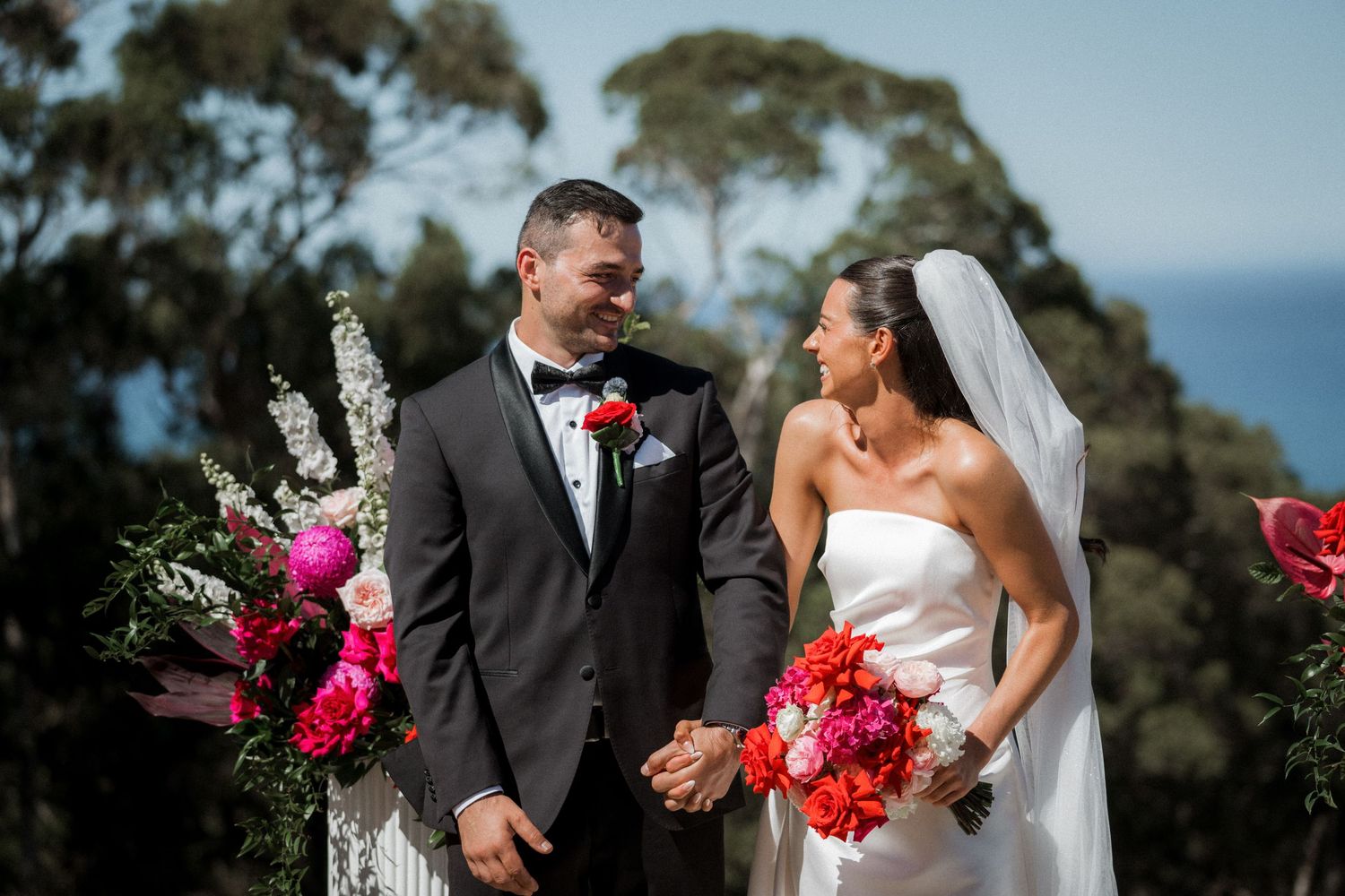 Wedding couple holding vibrant pink and red bouquets outdoors against a blue sky background.