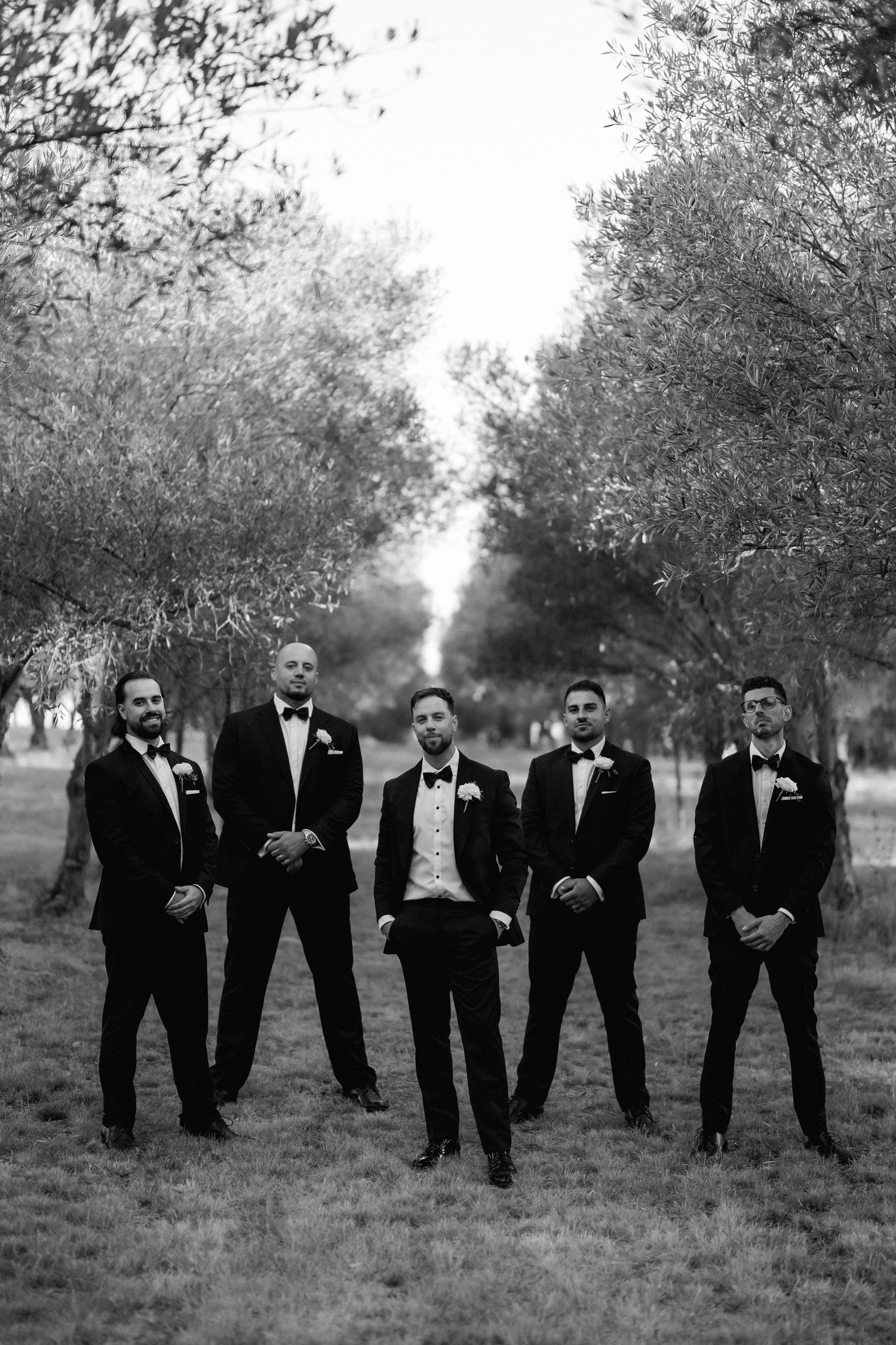 A group of groomsmen in black tuxedos pose together outdoors in a grassy field with trees in the background.