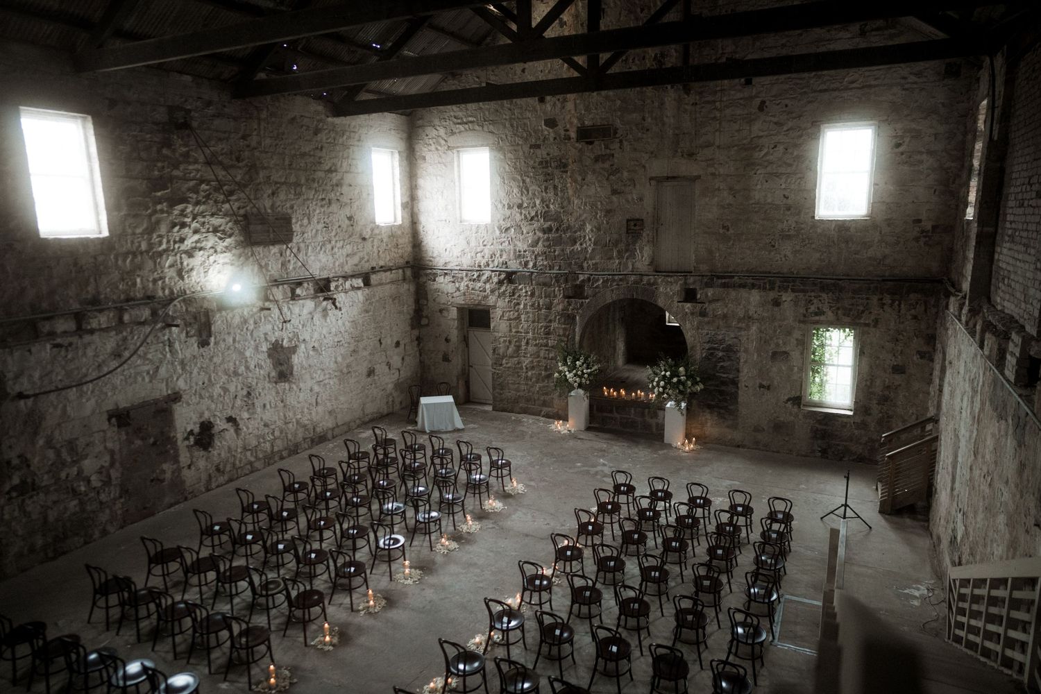 Rows of empty chairs arranged in a dimly lit stone room with natural light streaming through windows.