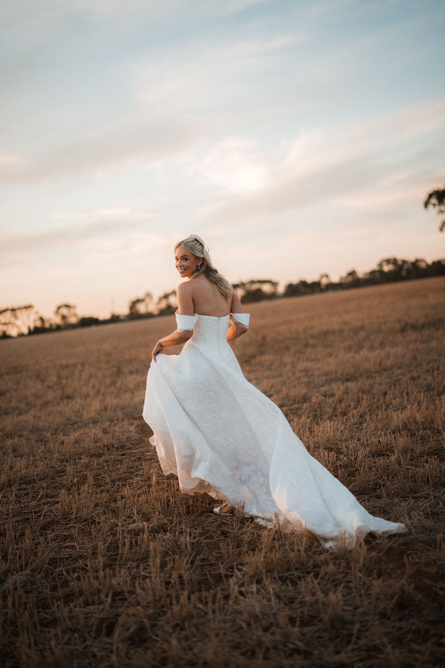 Bride in off-shoulder white lace gown twirls in a golden wheat field at sunset.