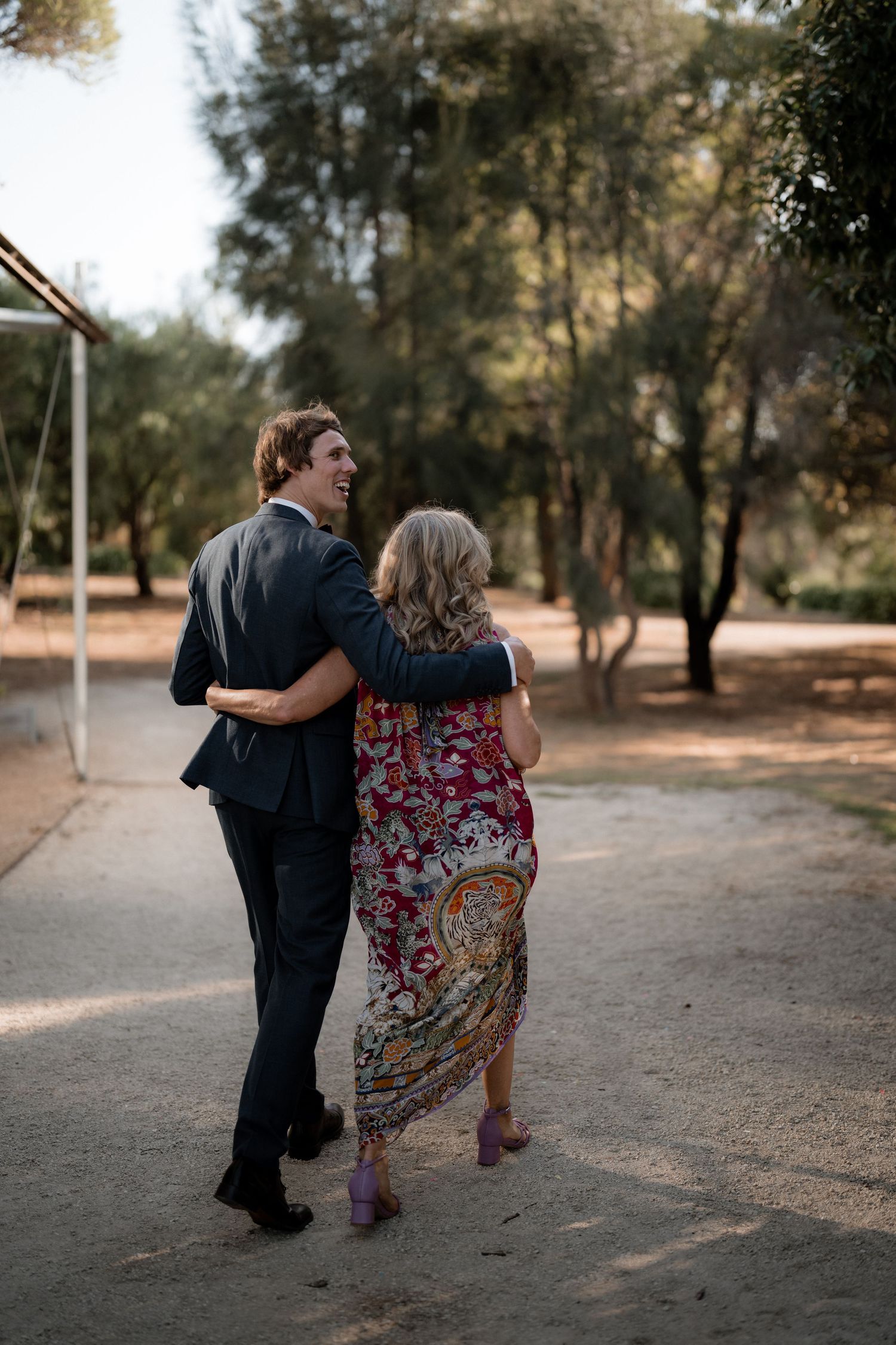 A couple walking arm in arm along a dirt path, with the person in a patterned maxi dress and another in formal attire.