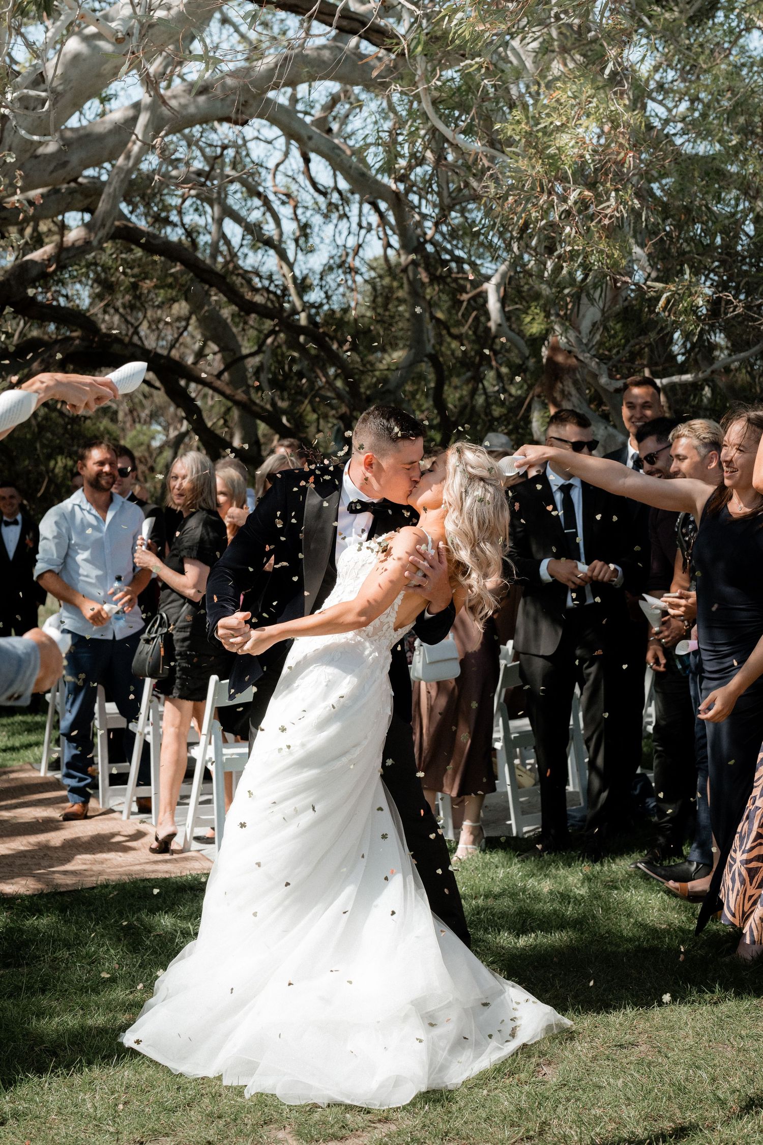 Newlyweds share their first dance surrounded by celebrating guests throwing confetti under a tree canopy during outdoor wedding.
