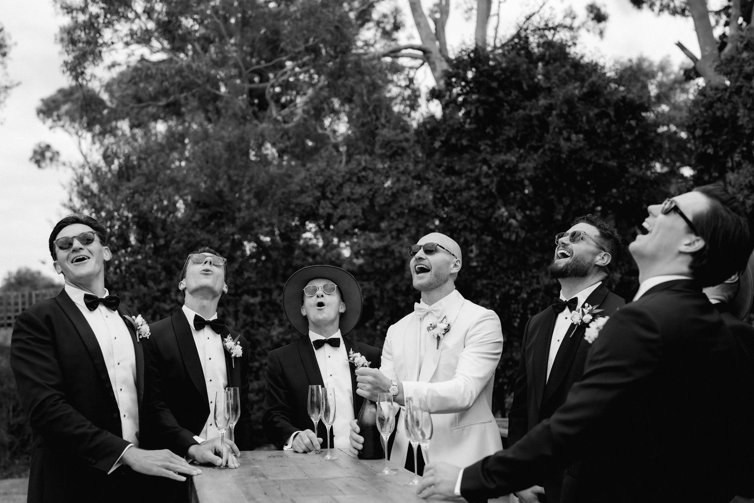 Black and white photo of groomsmen in tuxedos looking up and laughing during wedding celebration outdoors.