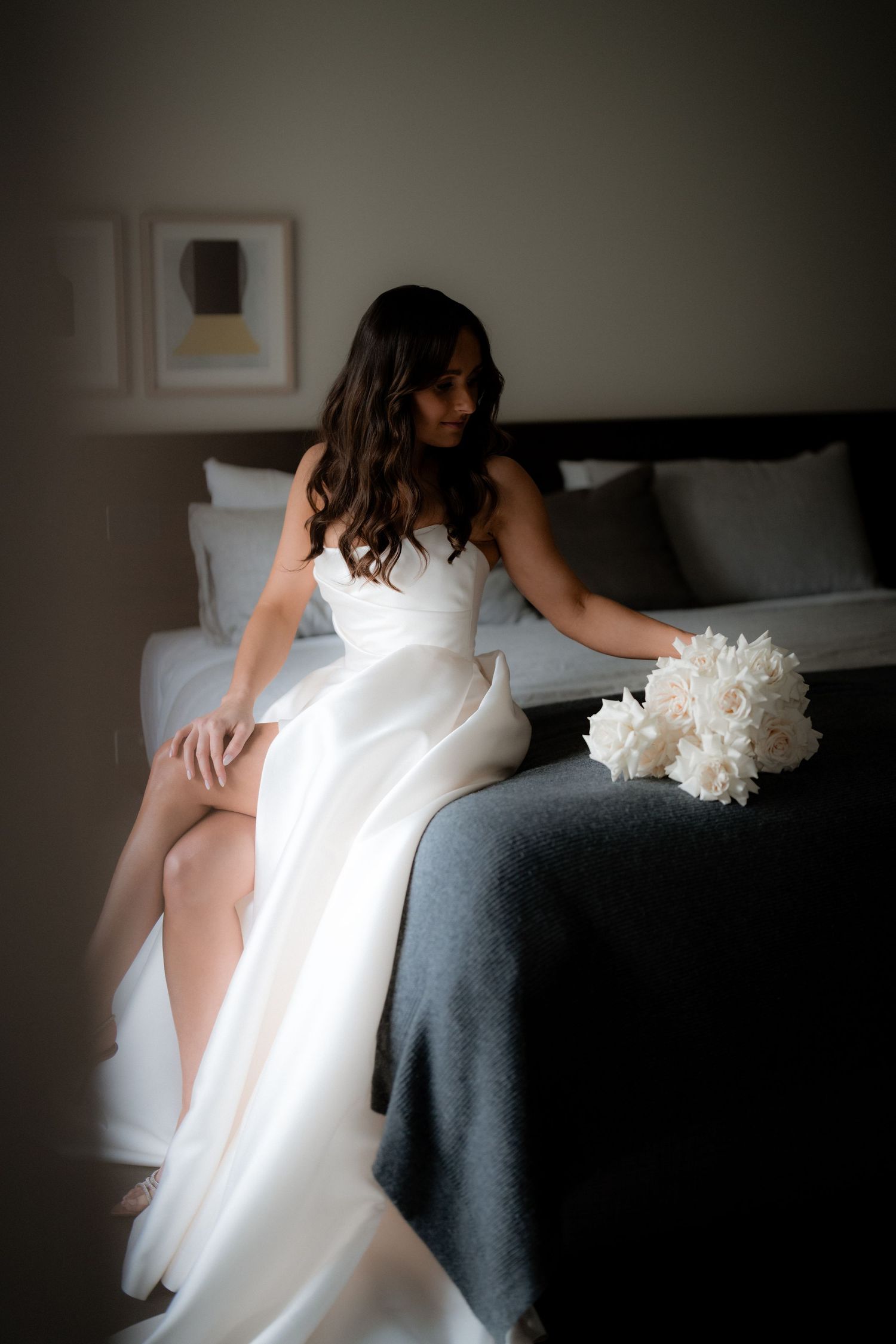 Bride in white gown sitting on grey couch with bouquet in moody lighting.