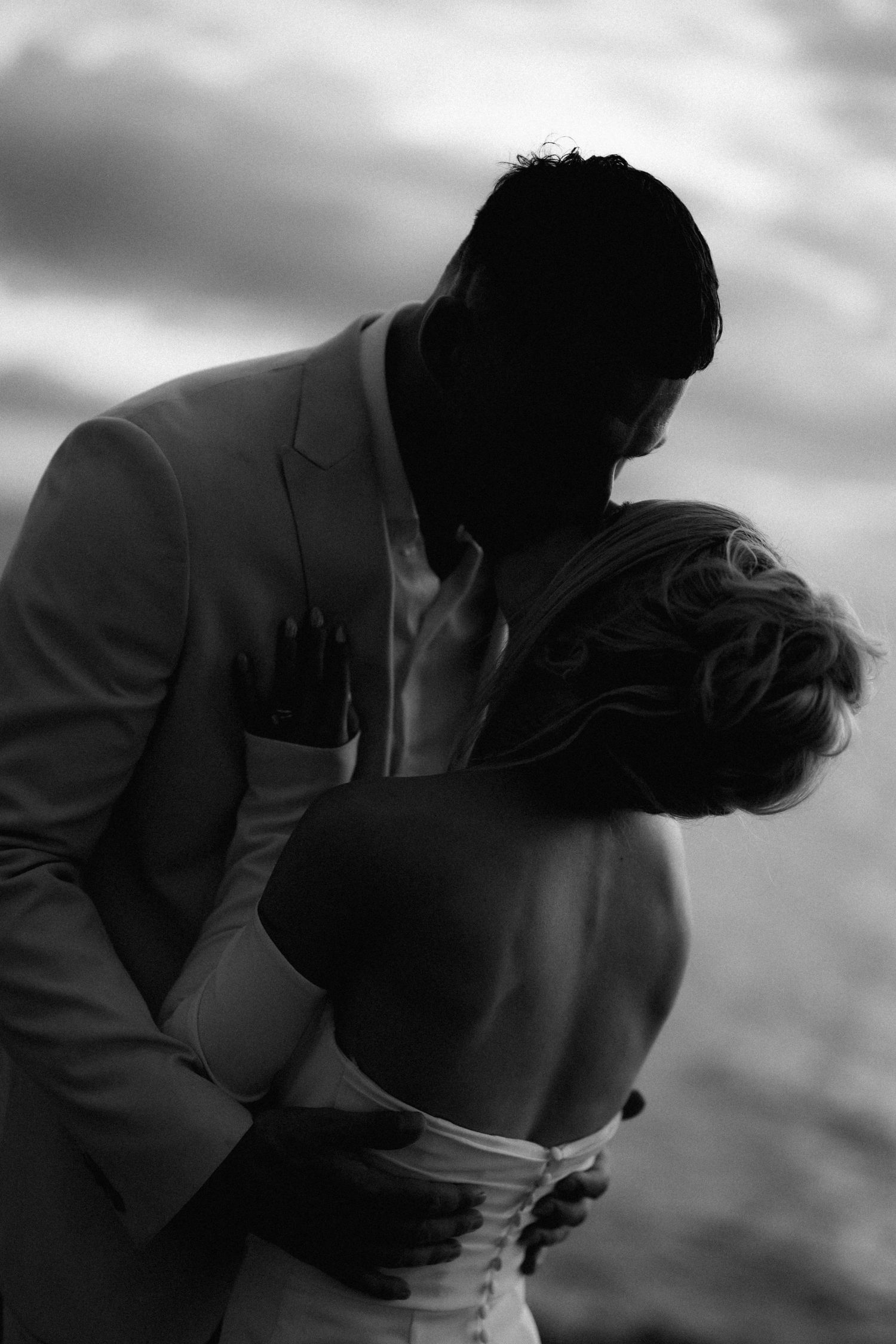 A dramatic silhouette of a bride and groom embracing against a moody sky in black and white.