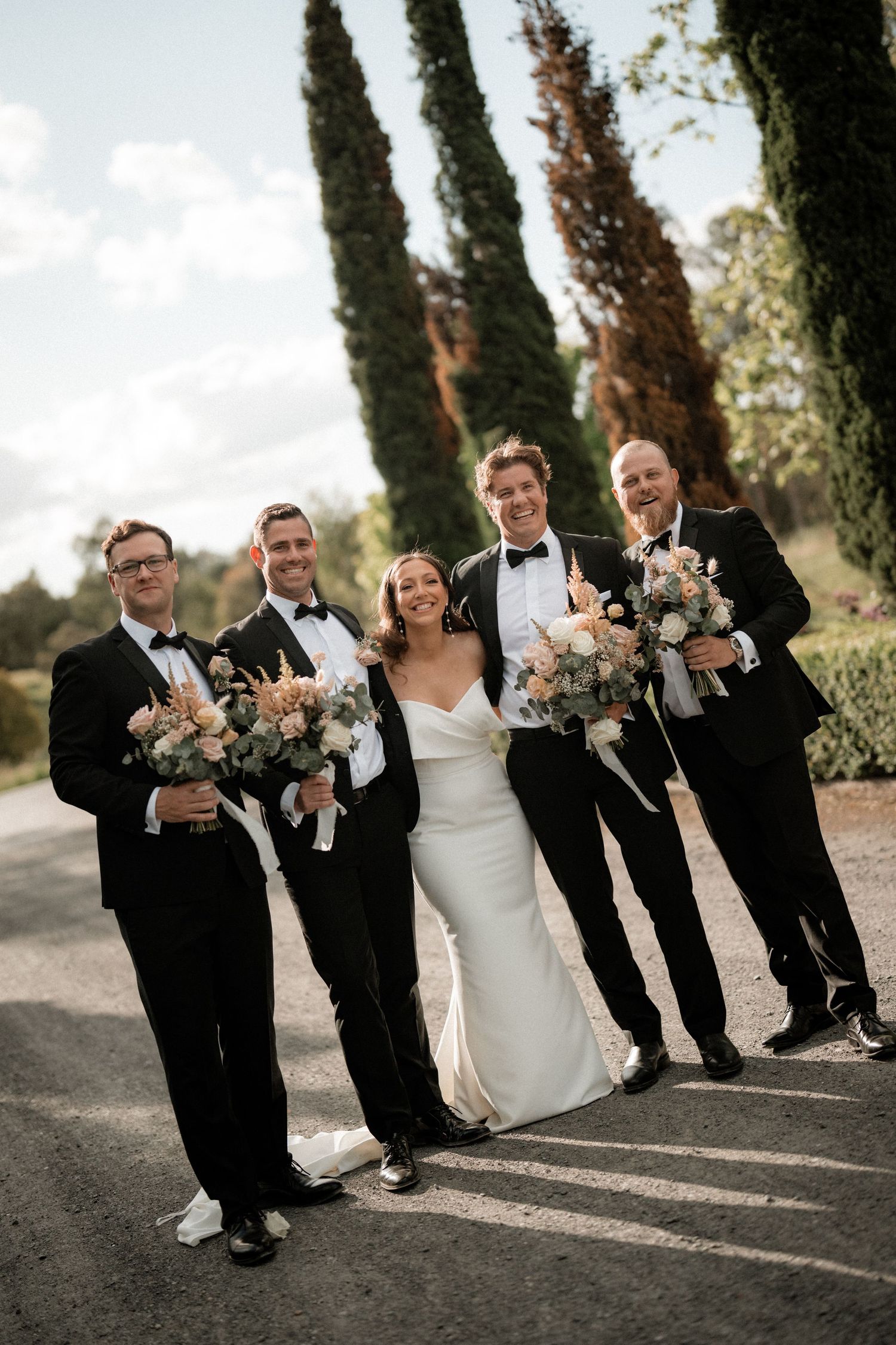 Wedding party strikes fun poses in formal black tuxedos and white dress against a backdrop of tall cypress trees.