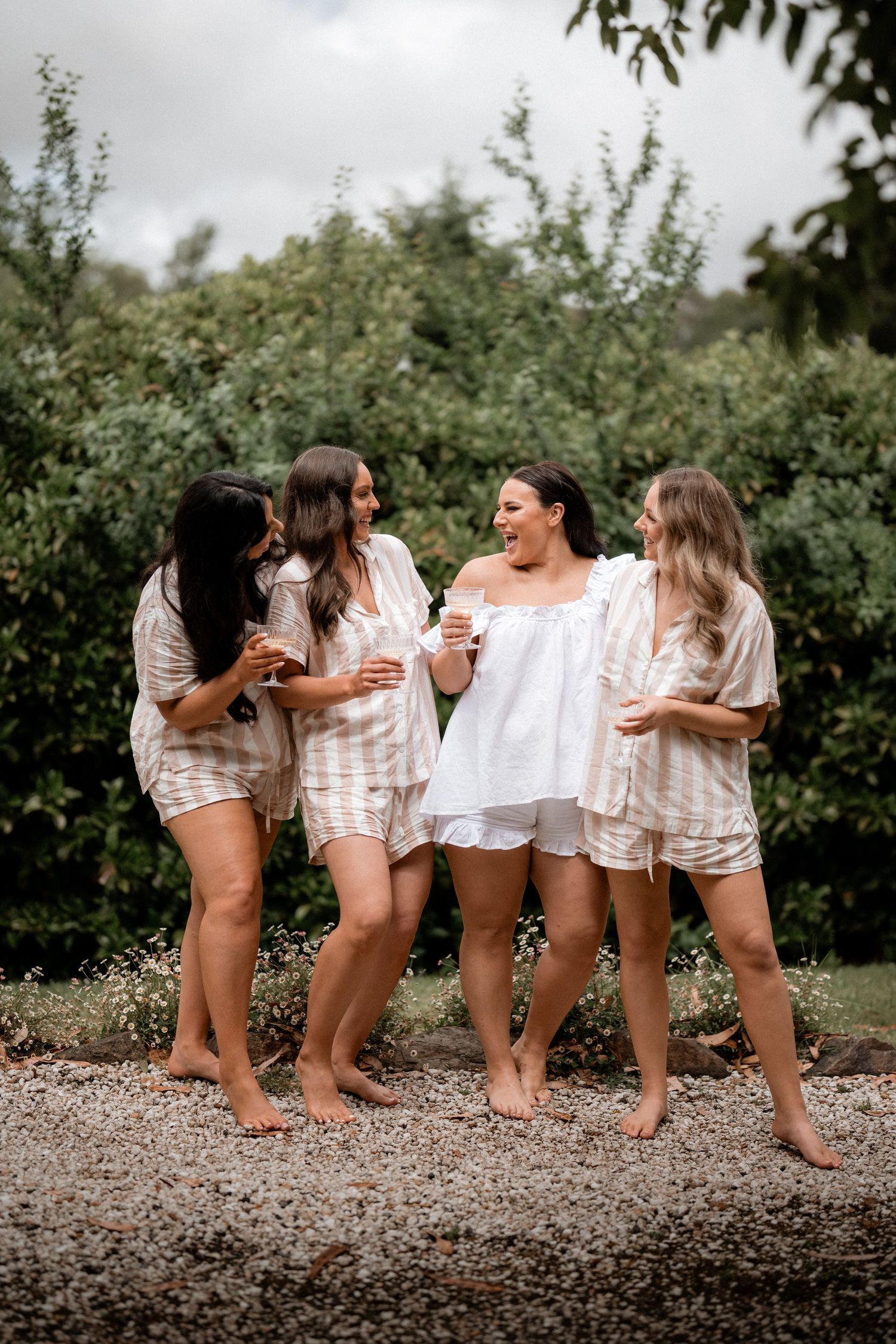 A group of friends in matching loungewear share laughter and drinks together on a gravel path surrounded by greenery.