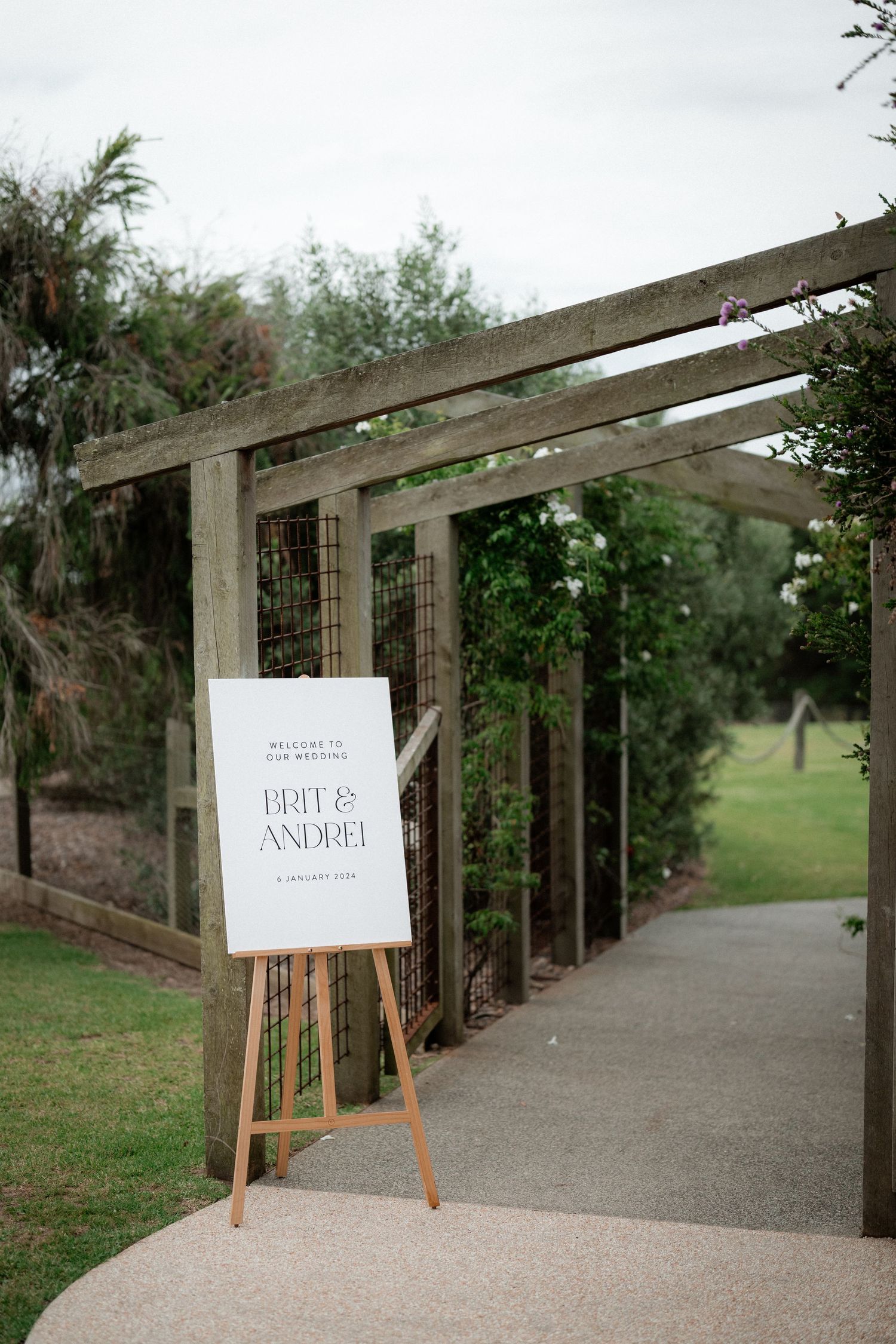 A wedding welcome sign on an easel stands beneath a wooden pergola along a garden pathway.