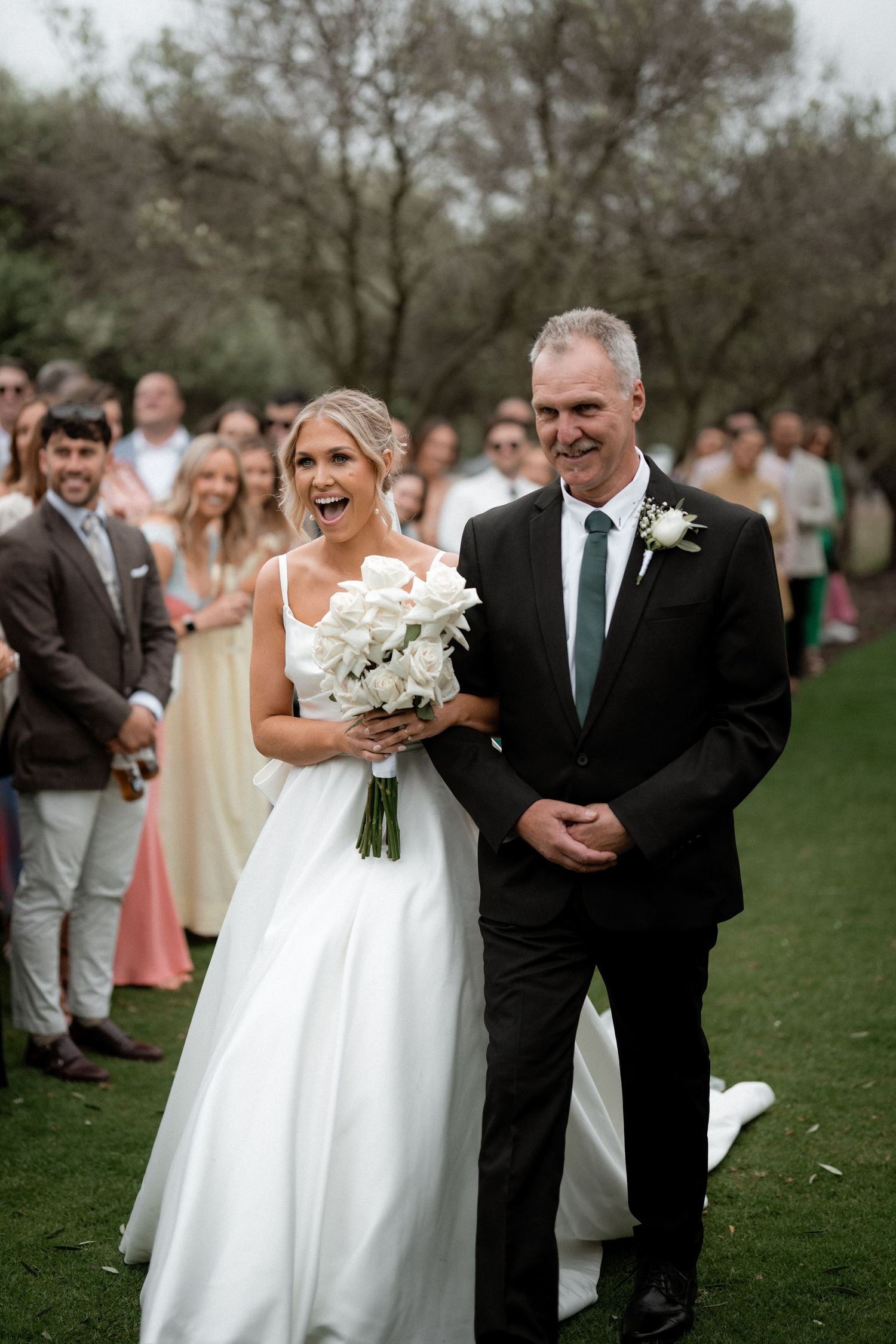 Father and bride walking down outdoor grassy aisle at wedding ceremony with natural setting and guests watching.