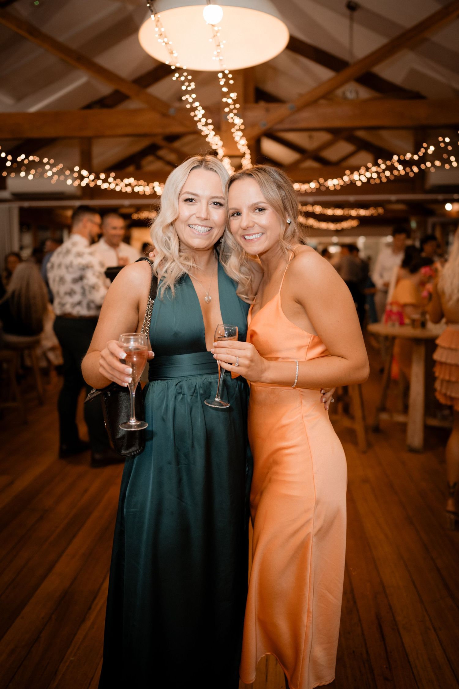 Wedding reception guests in formal attire enjoying drinks under warm string lights on a wooden dance floor.