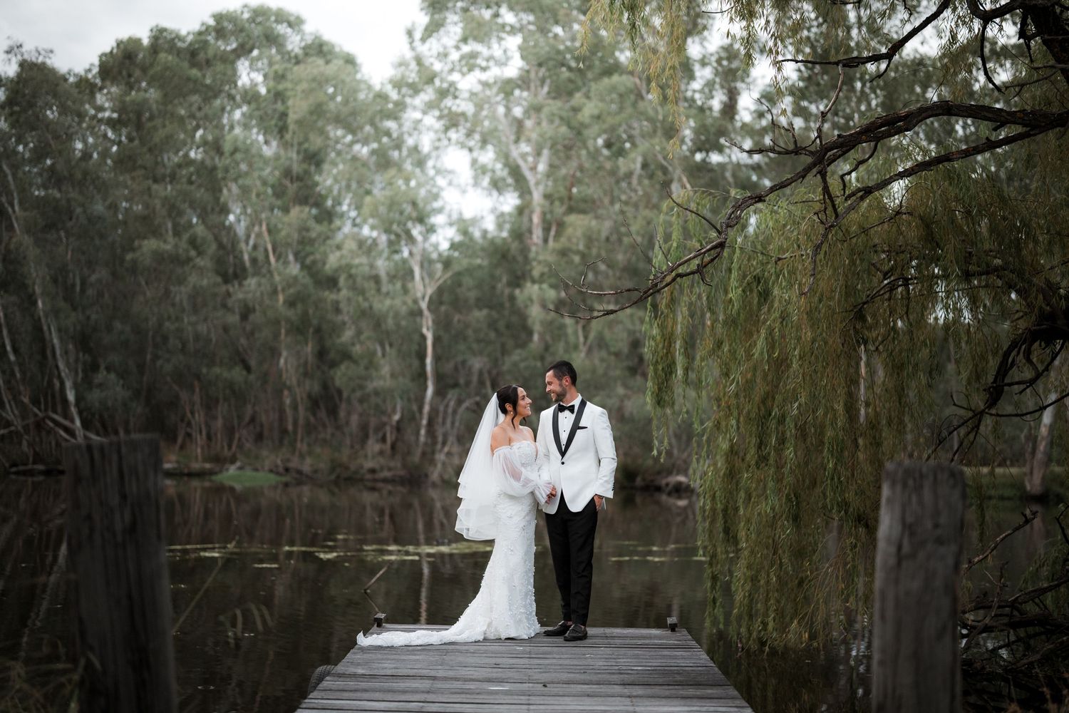 Scenic wedding photo shoot on a wooden bridge surrounded by willow trees and natural greenery.
