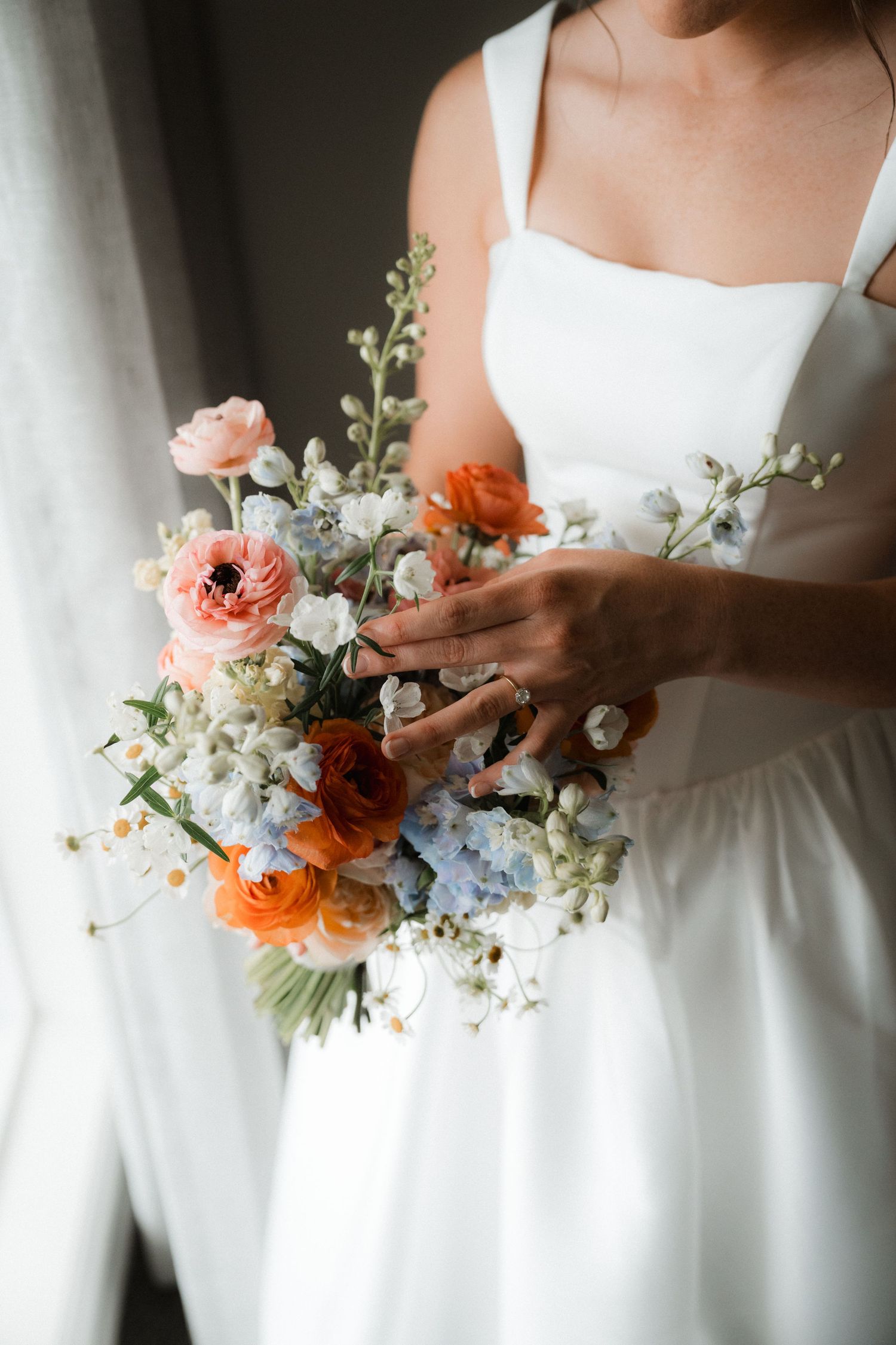 A delicate bridal bouquet featuring coral, white and blue flowers against a white wedding dress.