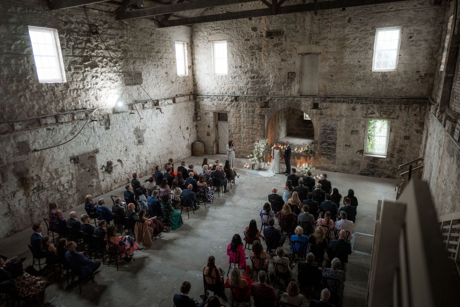 People gather inside a rustic stone building with high windows and exposed brick walls for what appears to be an event.