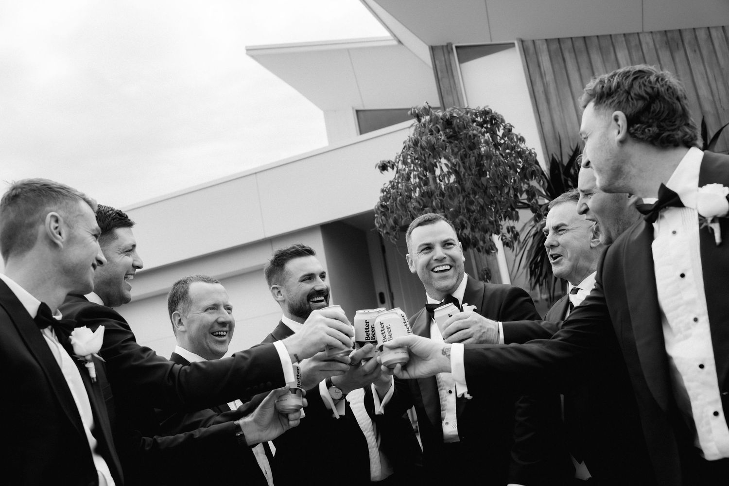 A black and white panoramic photo showing a group of men in tuxedos laughing and gesturing during a formal event.