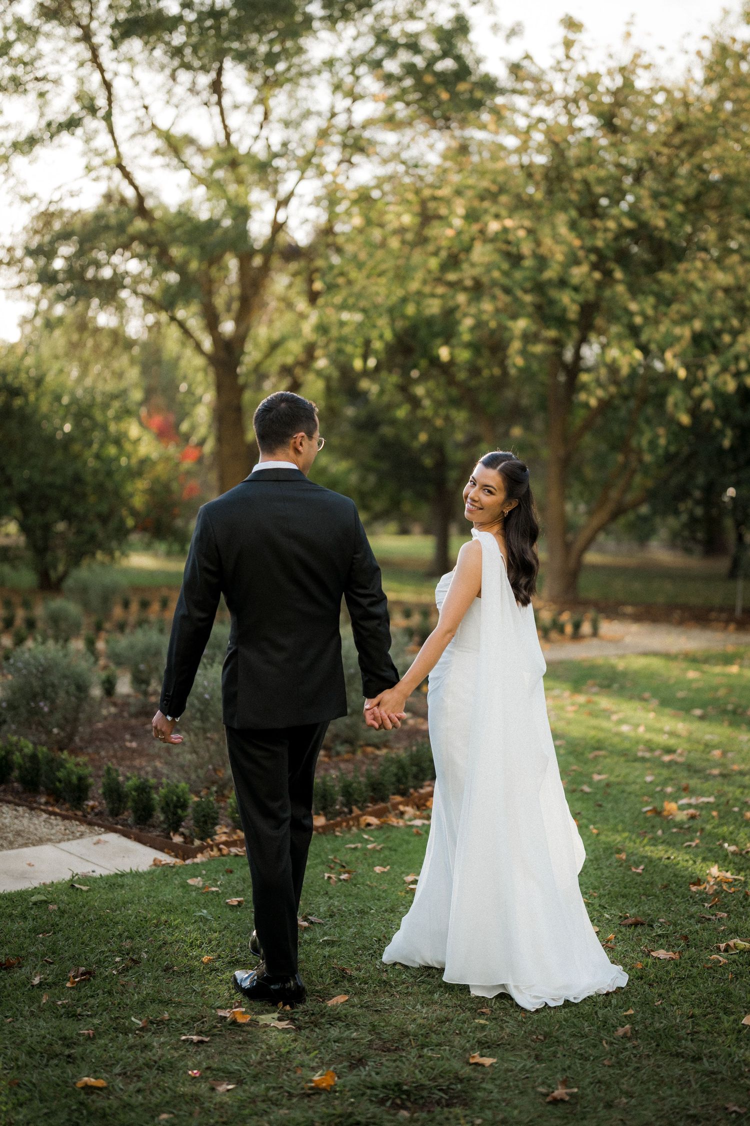 Bride in white gown and groom in black suit holding hands walking through lush garden.