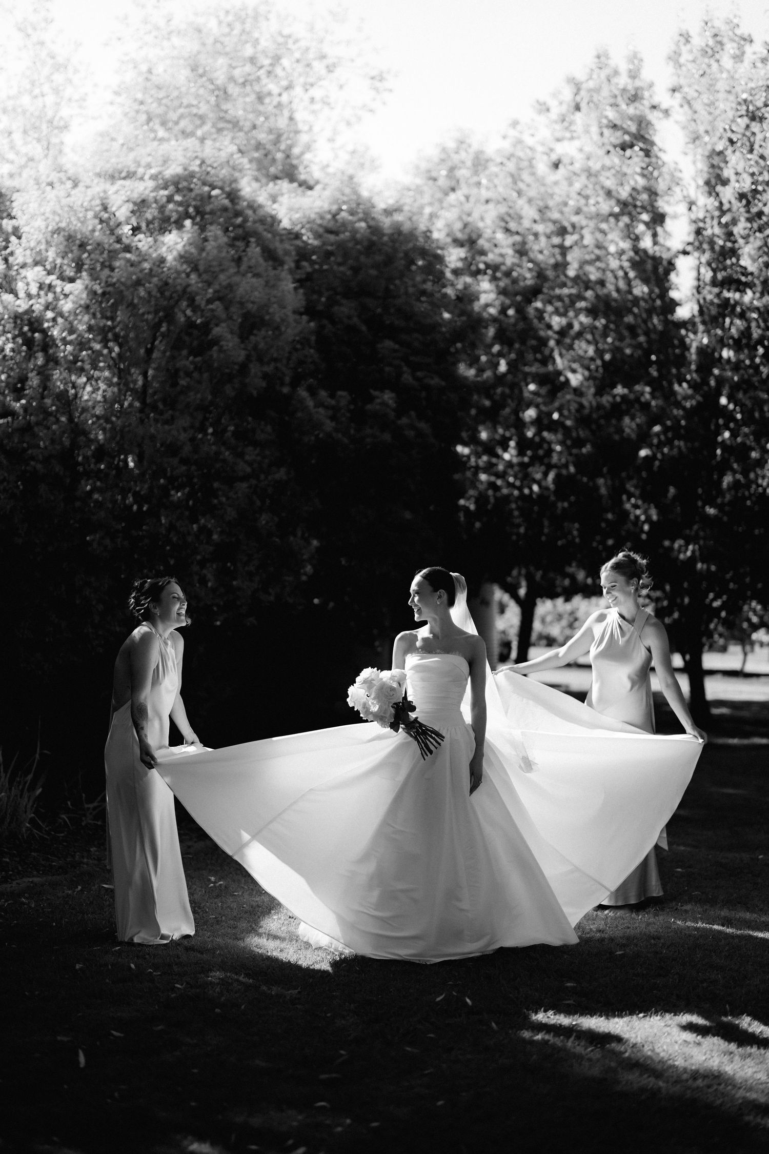 Artistic black and white photo of bride twirling her dress with attendants in garden setting.