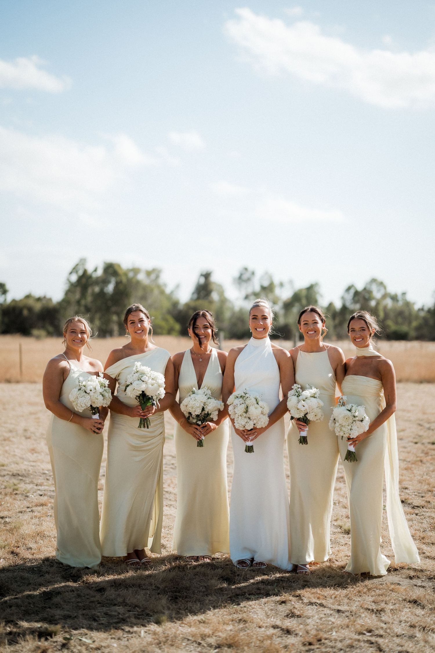 Elegant bridal party in cream-colored dresses holding white bouquets pose together in a sunlit outdoor setting.