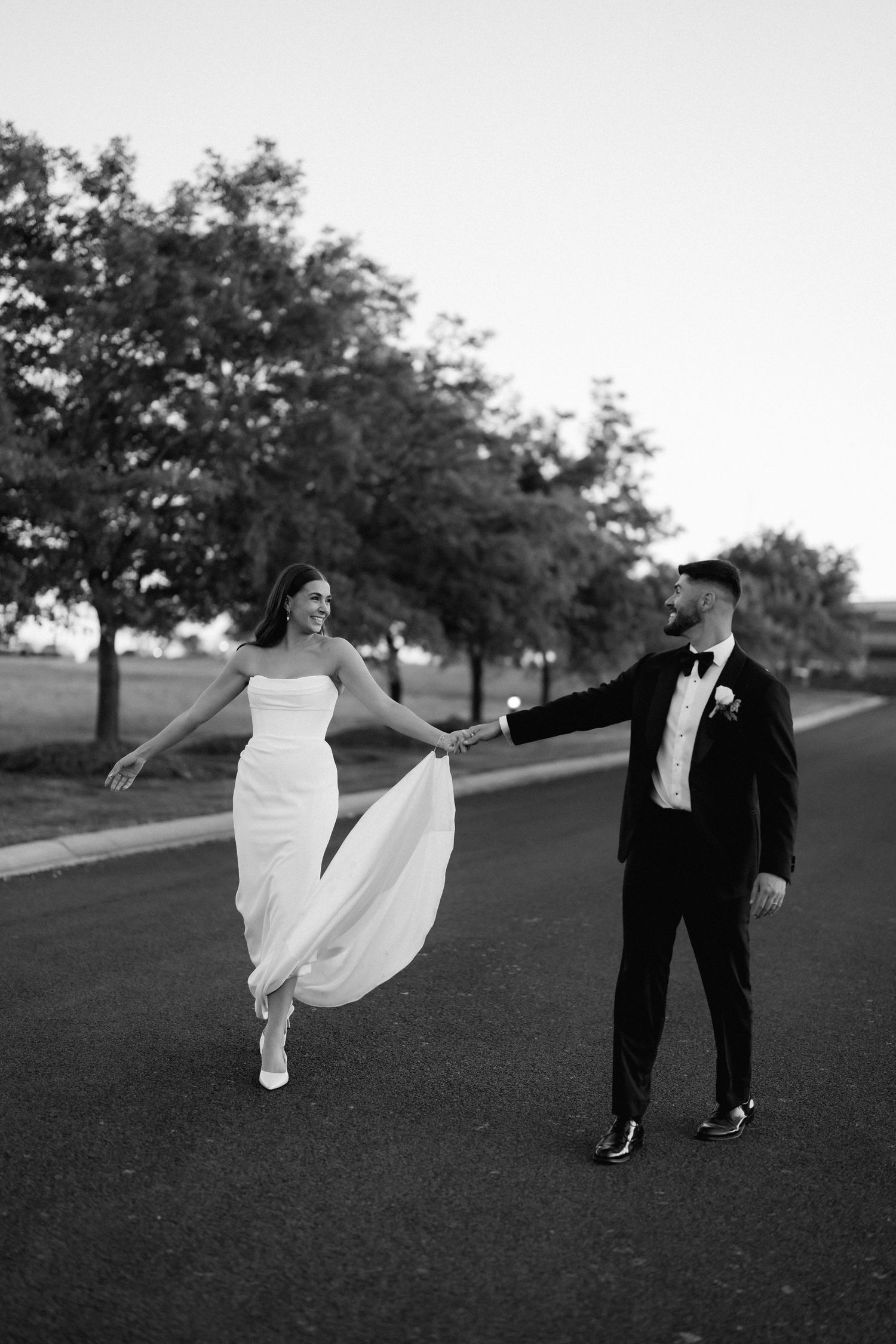 A couple in formal wedding attire playfully runs and spins across a grassy hill at sunset.