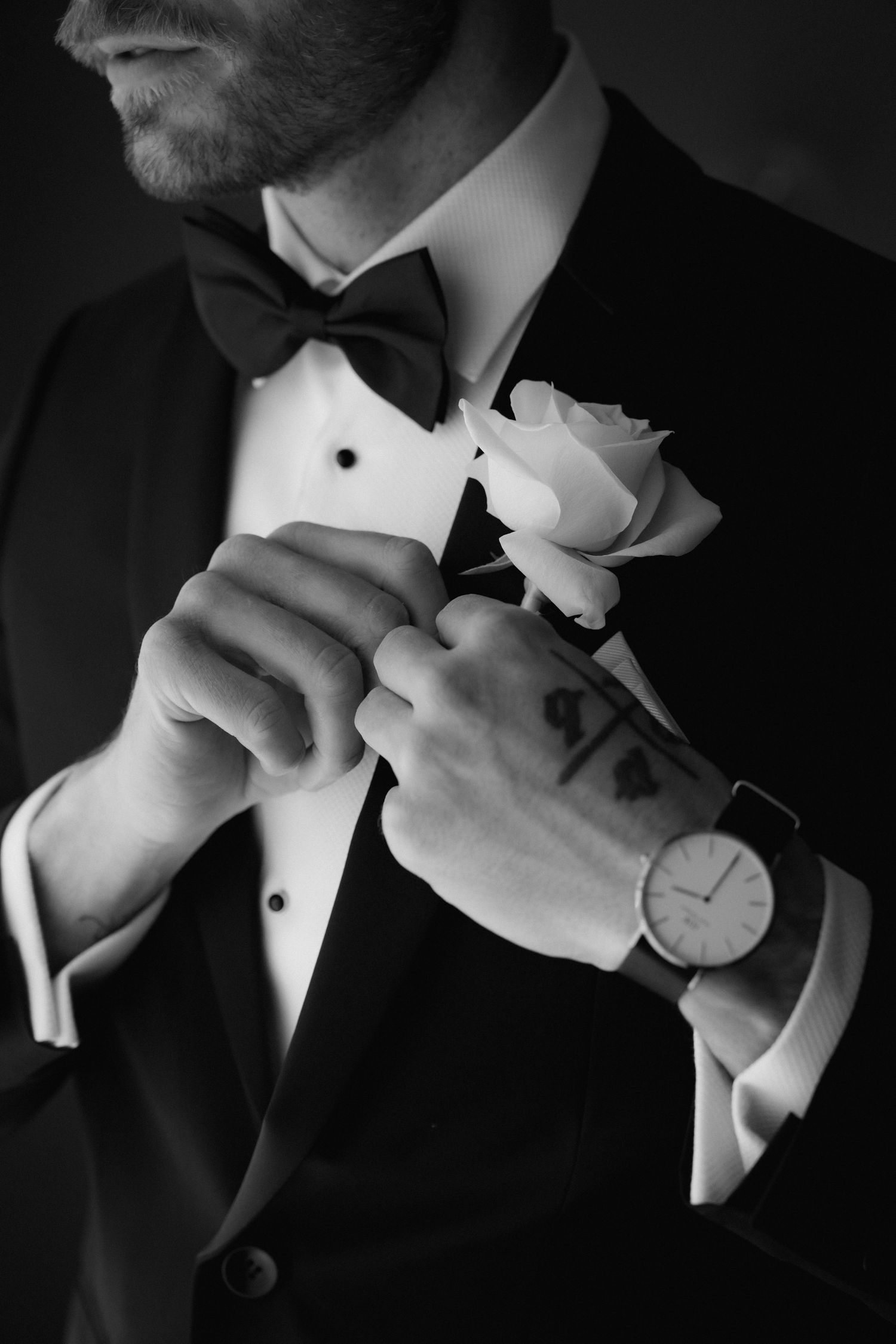 Close-up of groom in tuxedo adjusting white boutonniere flower with watch visible.