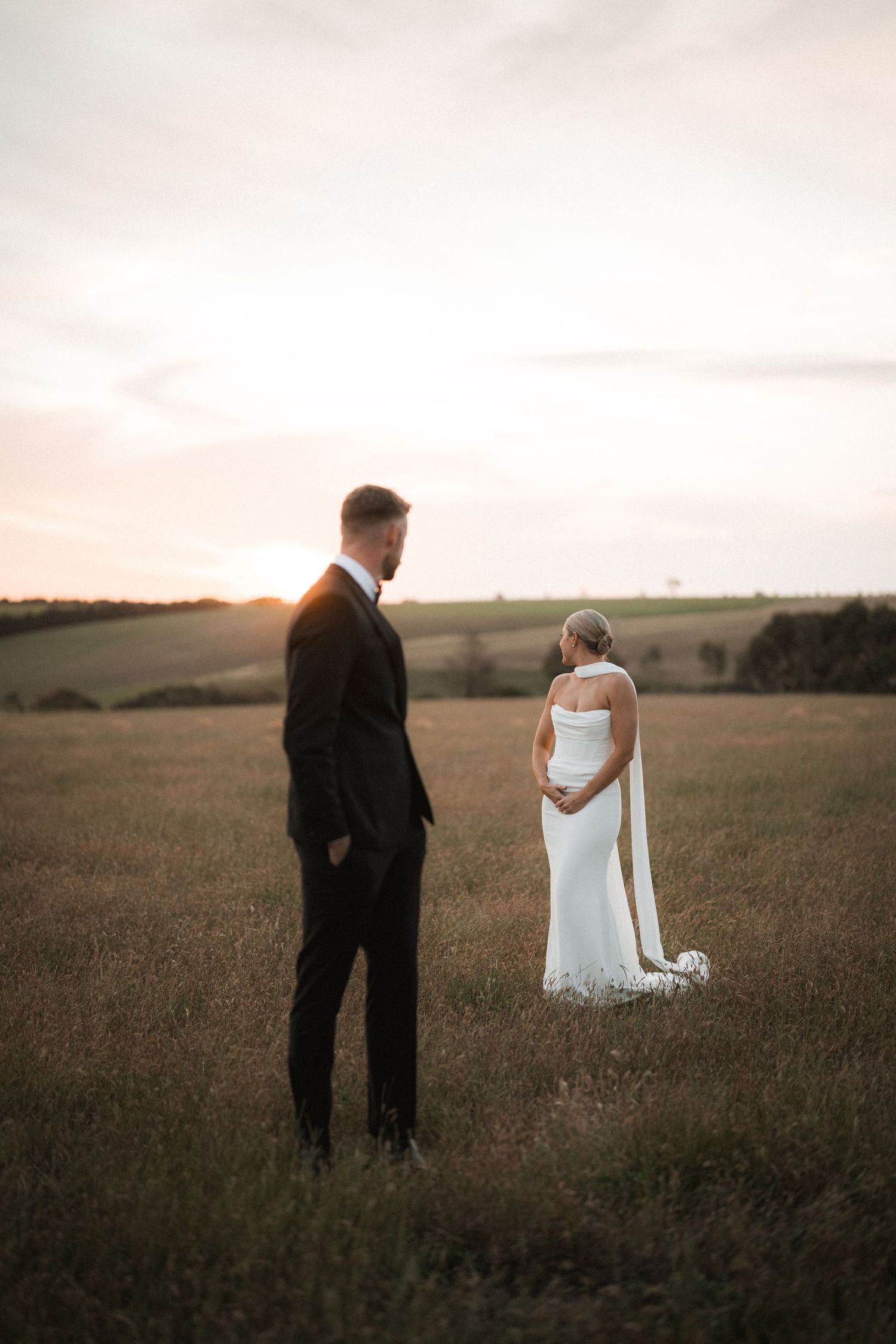 A couple in wedding attire stands together in a golden field at sunset, creating a romantic silhouette against the horizon.