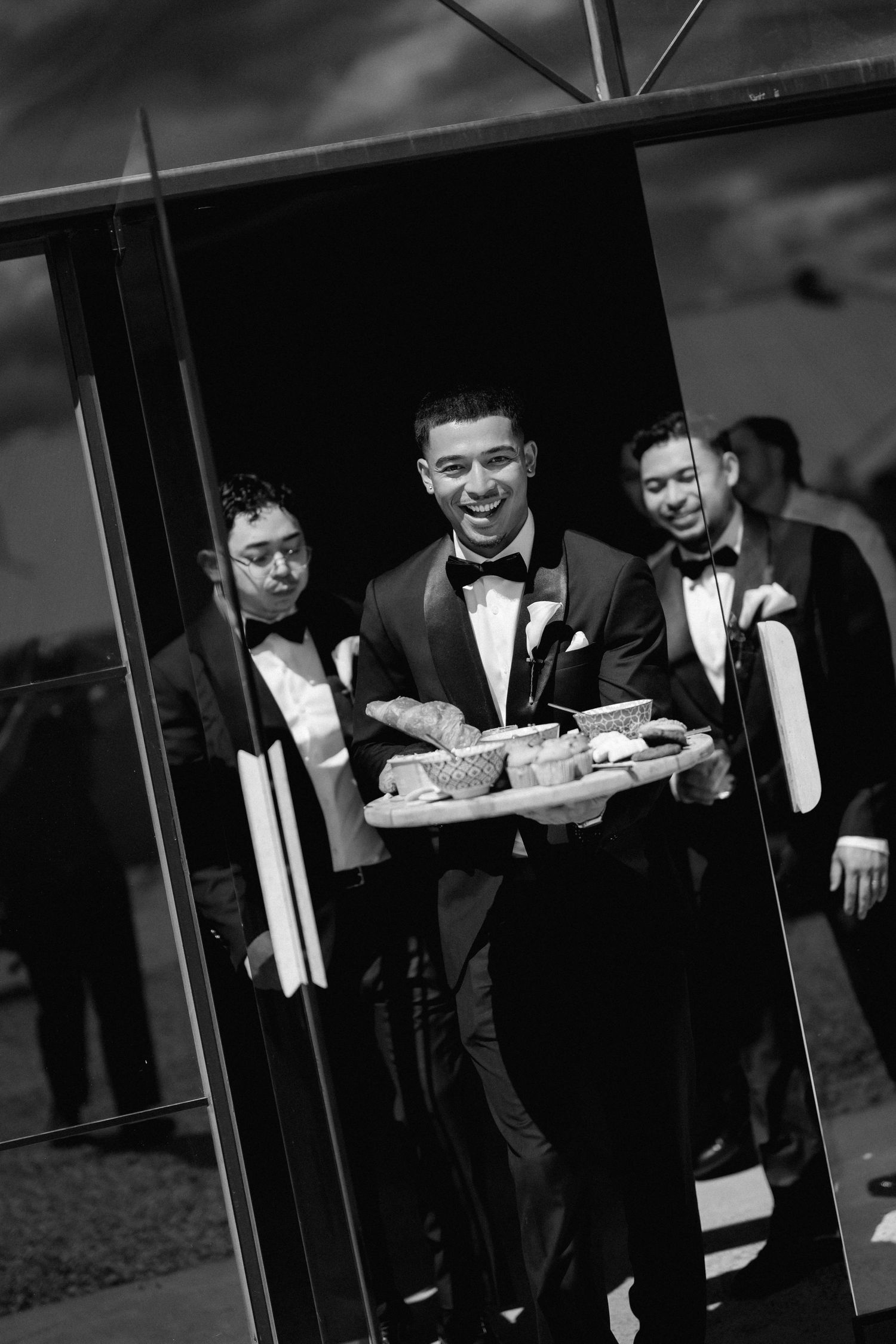 Smiling groomsman in a black bow tie carries a tray of snacks past fellow groomsmen in a black and white wedding photo.