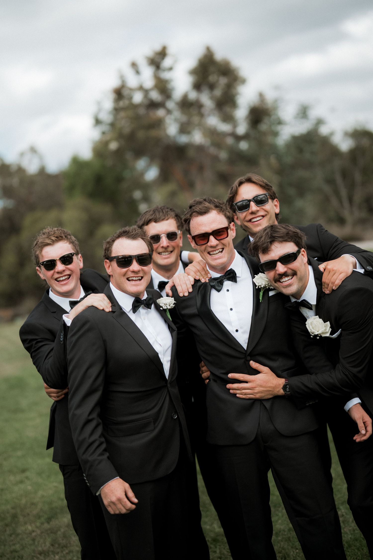 A group of groomsmen in black tuxedos and sunglasses share a candid laughing moment outdoors.