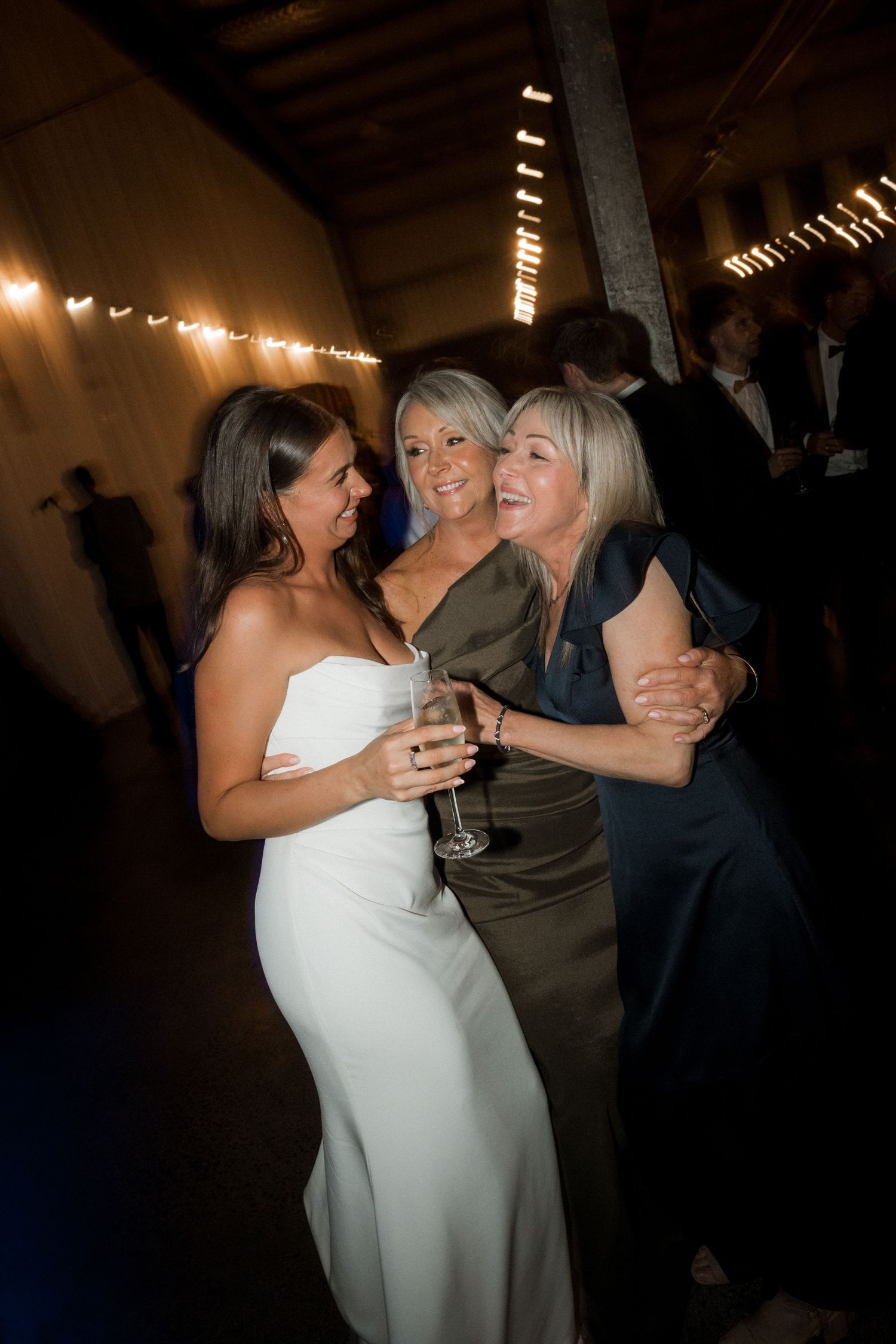 People dancing and embracing at a dimly lit wedding reception with string lights overhead.