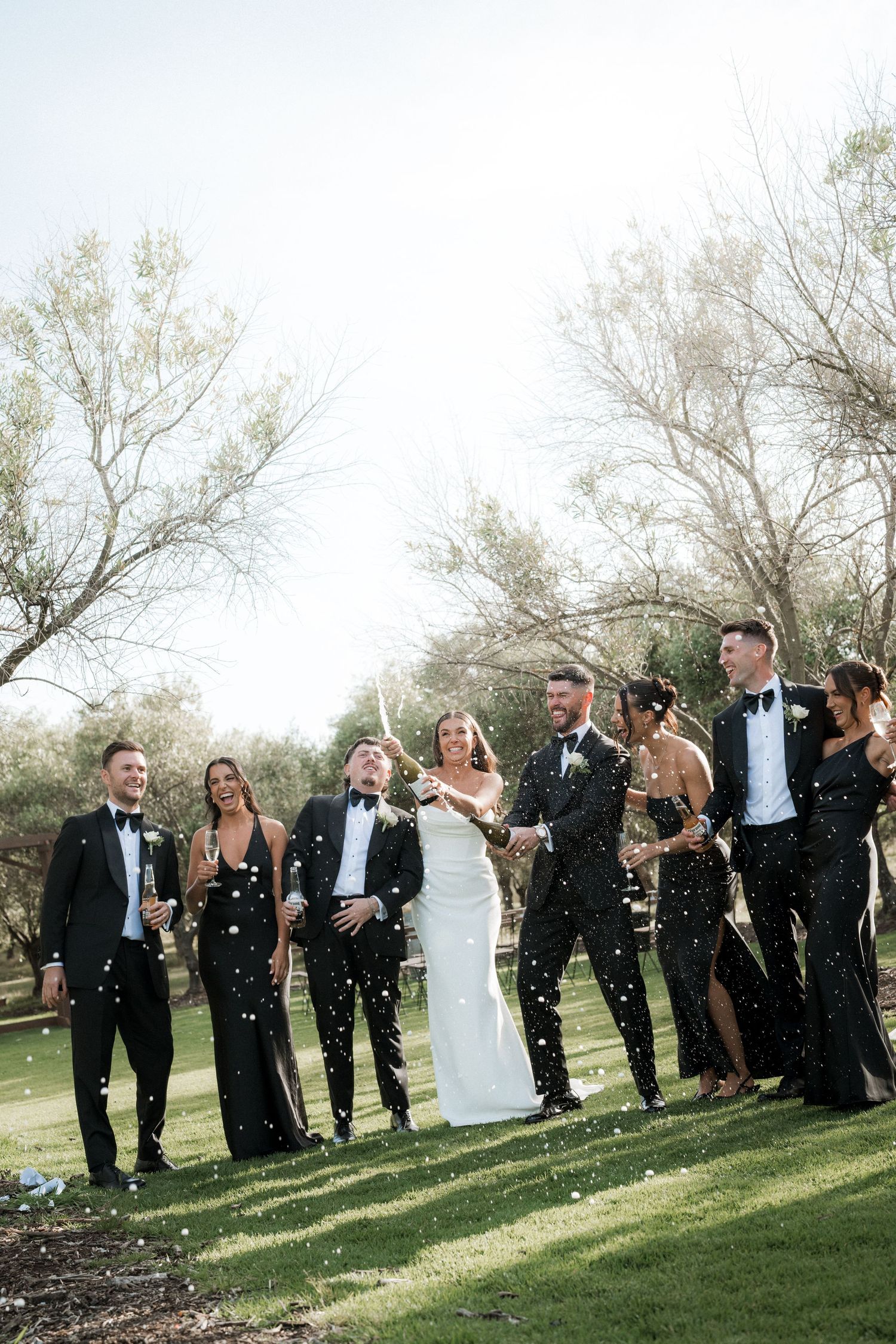 Wedding party celebrates outdoors with confetti under trees on a sunny day.