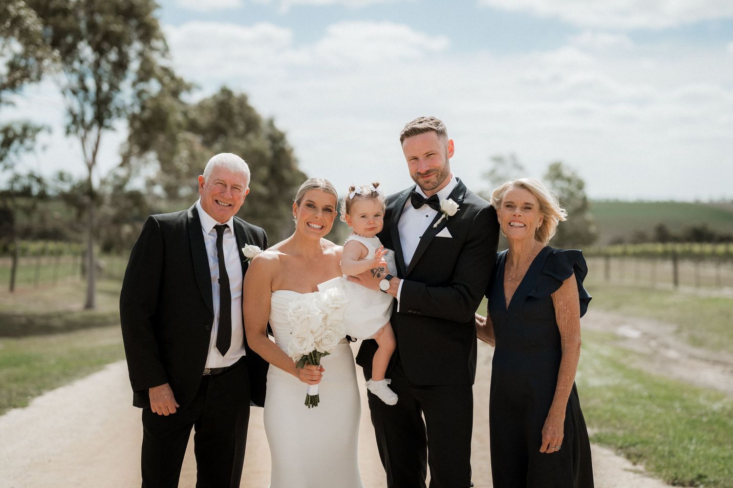 Wedding party poses together outdoors on a sunny day with rural landscape in background.