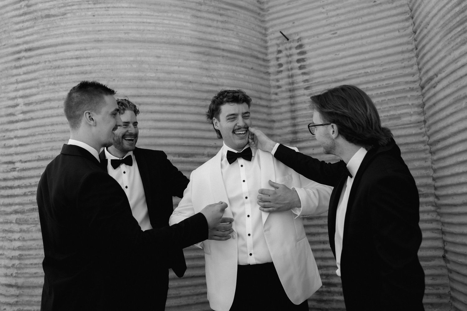 Black and white photo of groom in white jacket laughing with three groomsmen in tuxedos.