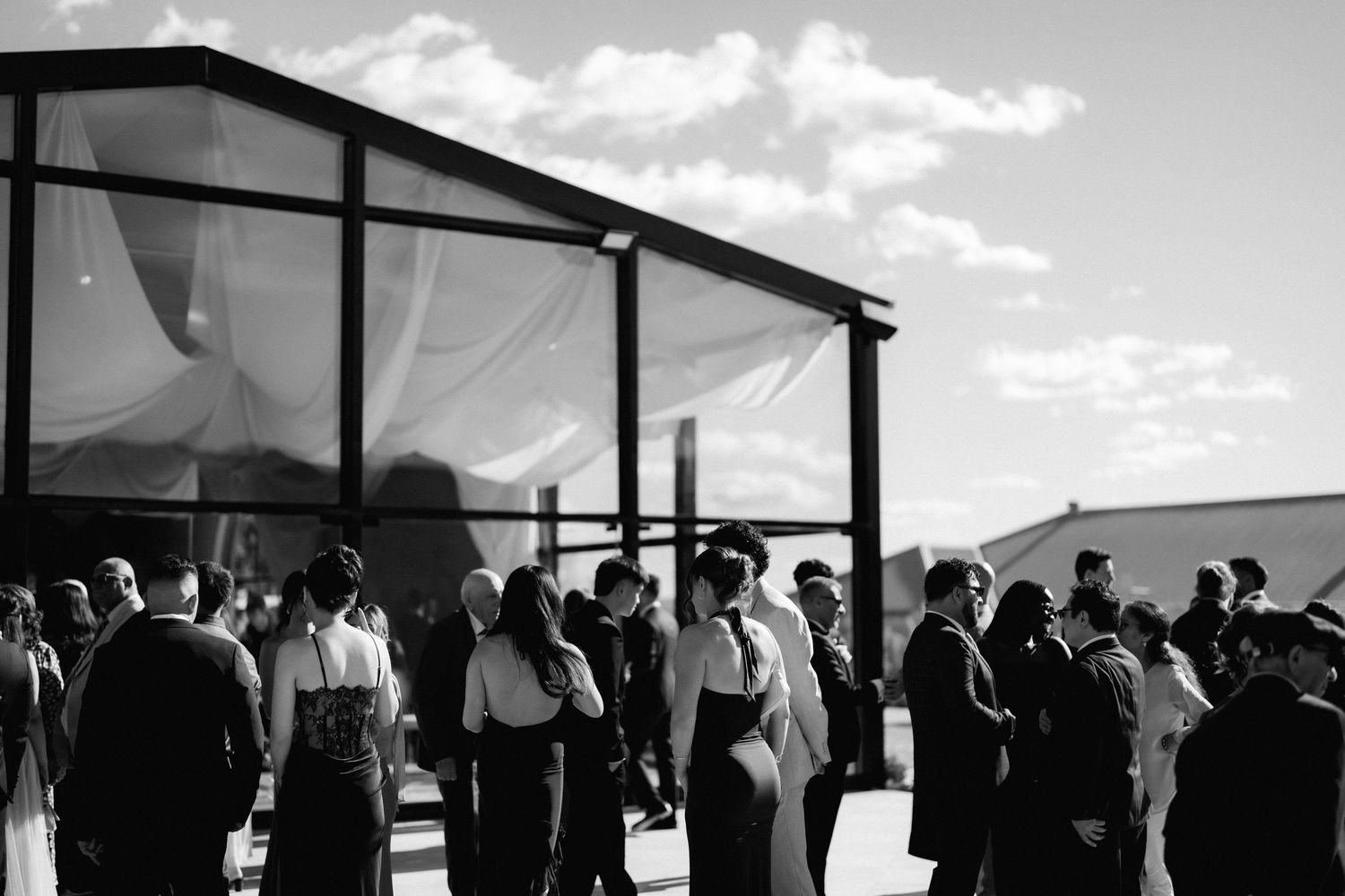 Black and white photo of formally dressed wedding guests gathered outdoors beside a draped open-air ceremony tent under a cloudy sky.