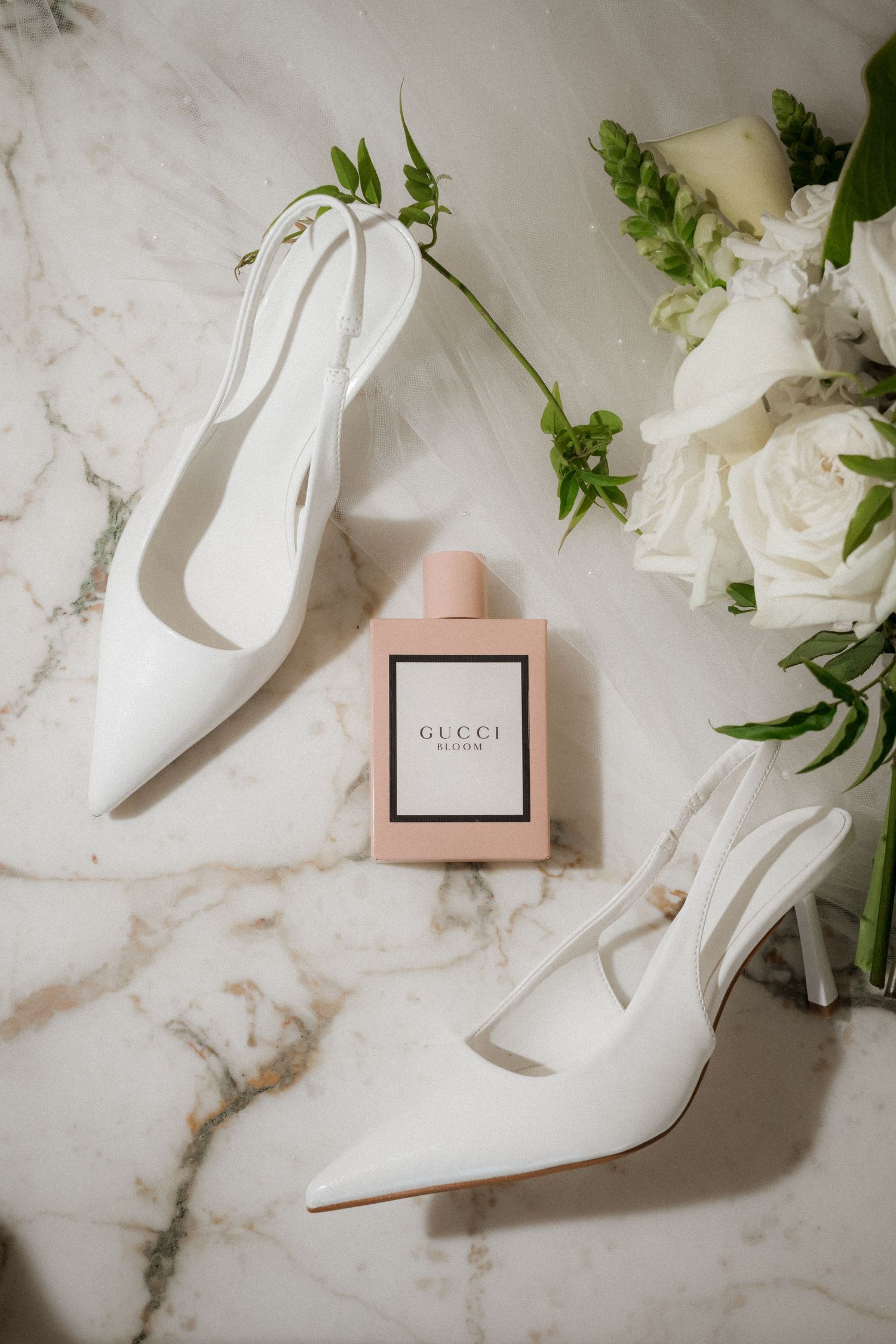 White pointed-toe slingback heels beside a Gucci Bloom perfume bottle and white rose bouquet on a marble surface.