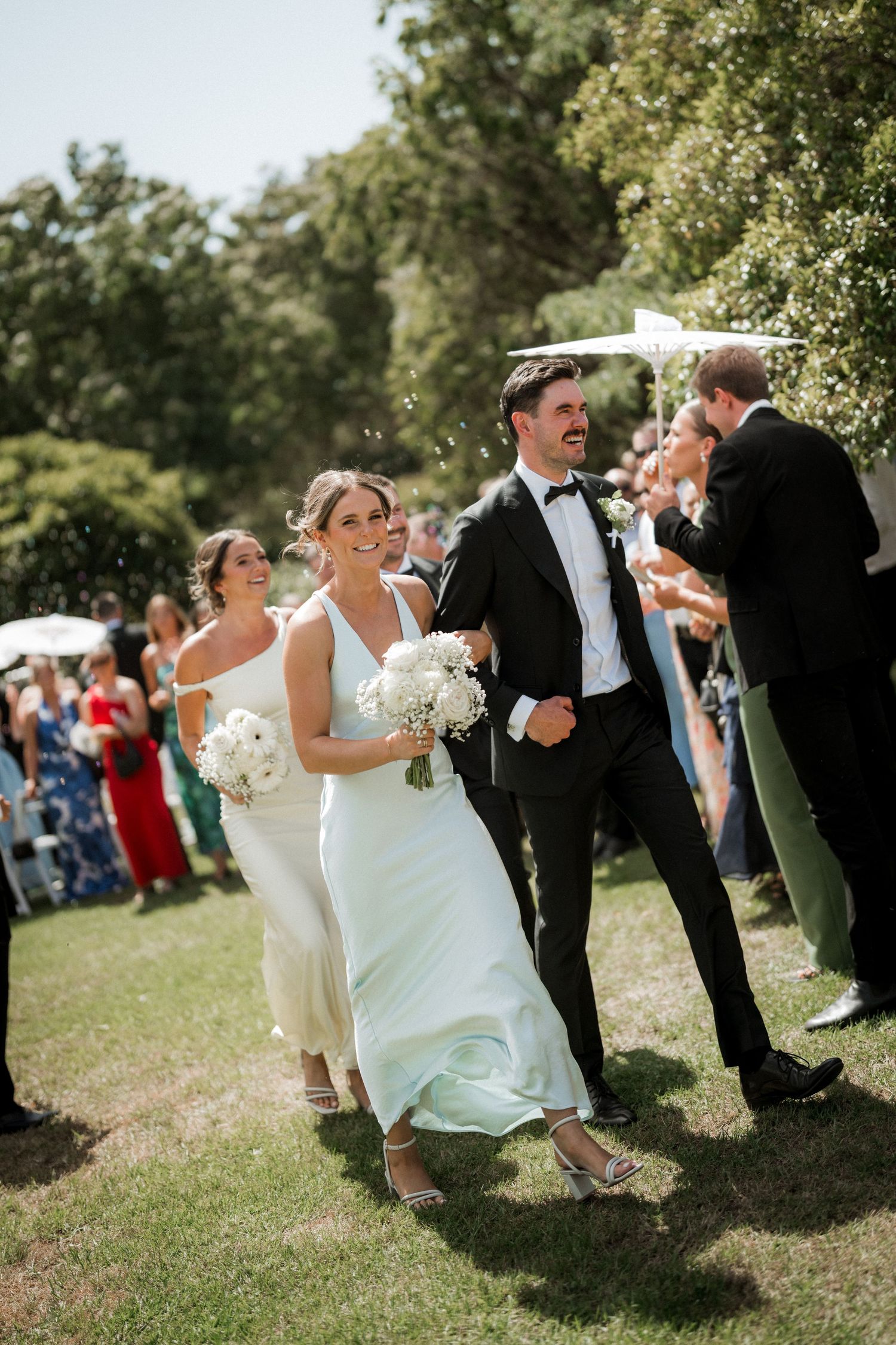 Wedding party celebrates on sunny lawn with guests in formal attire.