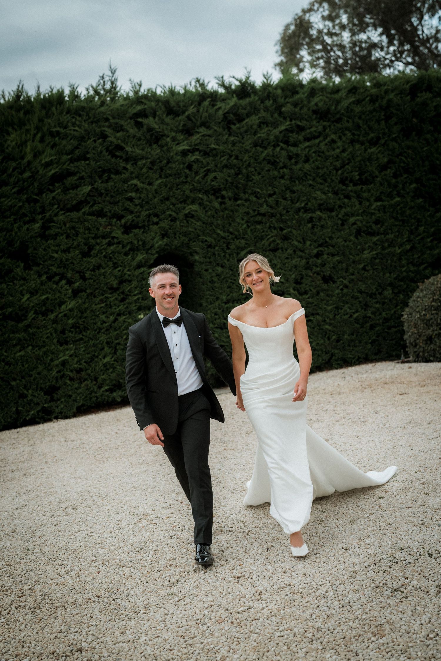 A couple in wedding attire walks playfully along a gravel path bordered by tall green hedges.