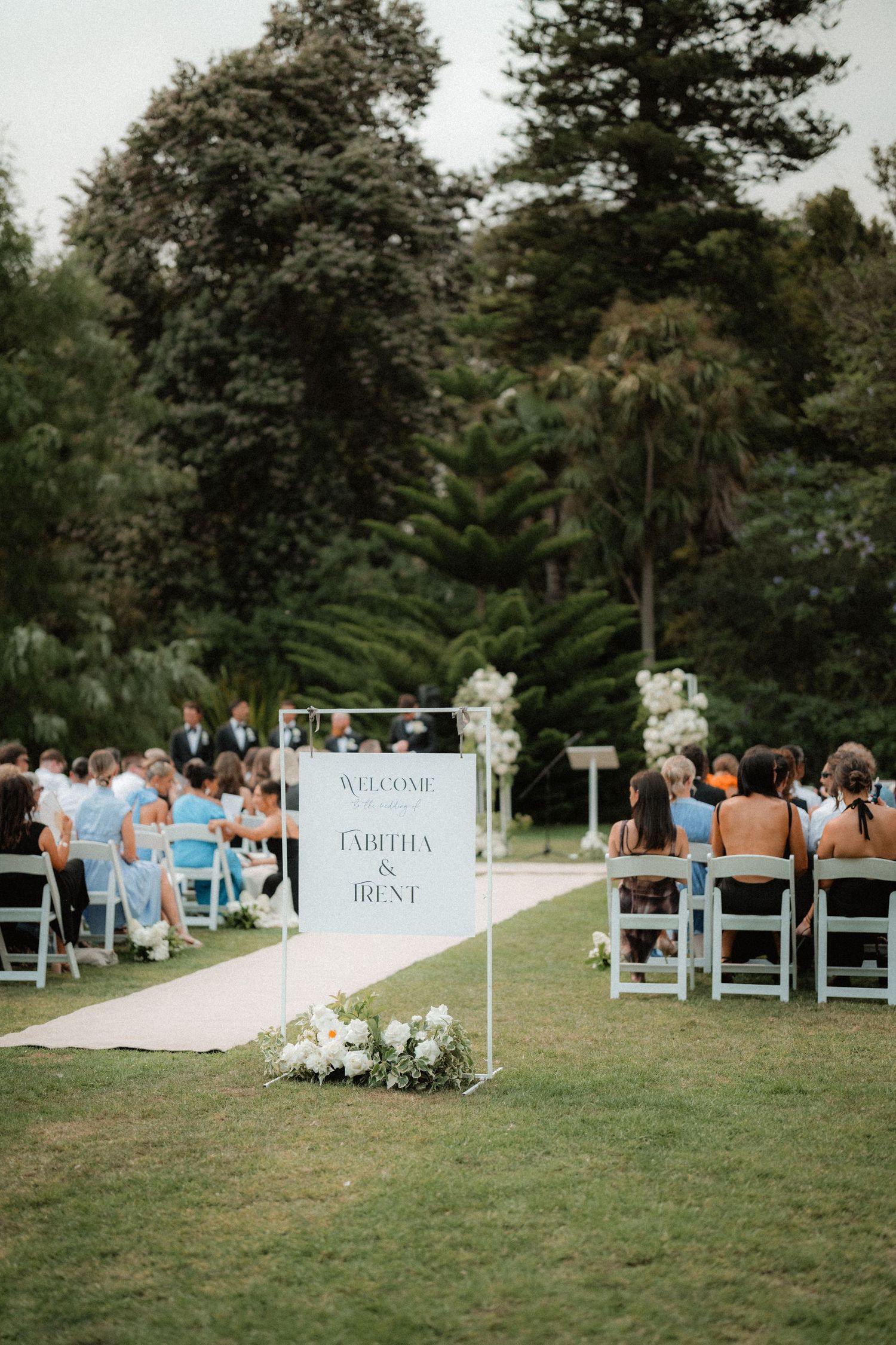 An outdoor wedding ceremony setup with white chairs and floral decorations along a white aisle runner surrounded by trees.