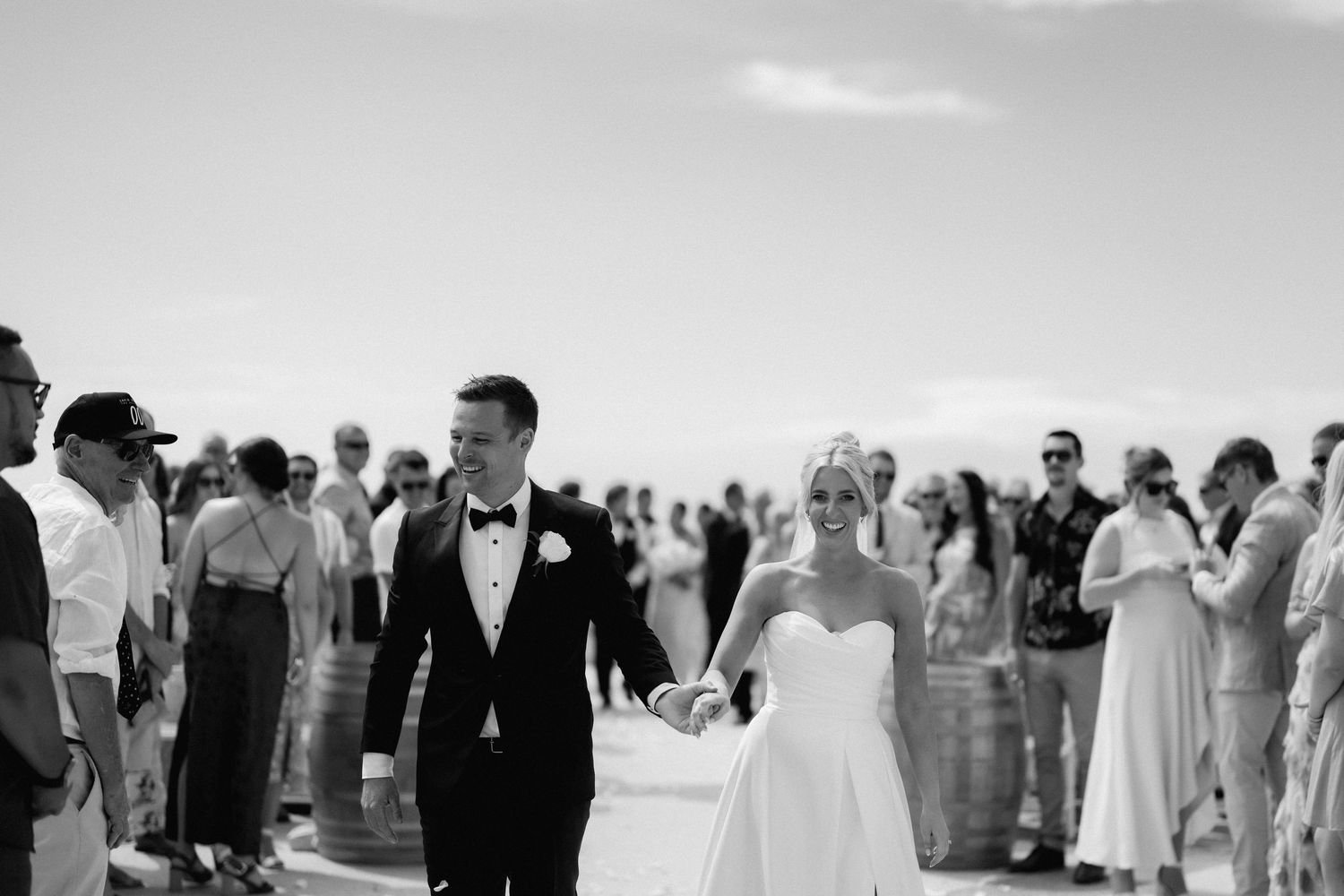 A black and white photo of newlyweds walking among wedding guests at an outdoor ceremony.